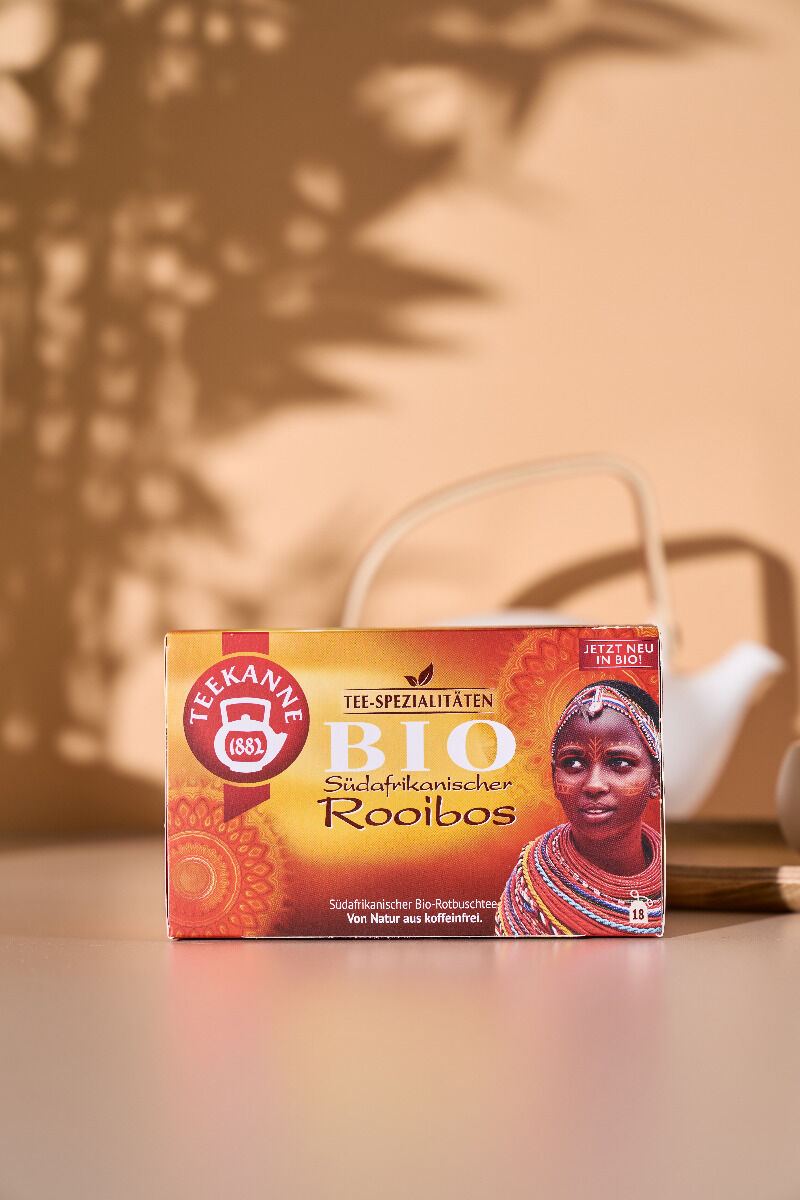 Eine Schachtel Südafrikanischer BIO Rooibos RFA-Tee steht auf einem Tisch und zeigt das Bild einer Frau in traditioneller Kleidung. Im Hintergrund erscheint eine unscharfe weiße Teekanne. Warmes Licht und Schatten von Blättern werfen Muster an die Wand.