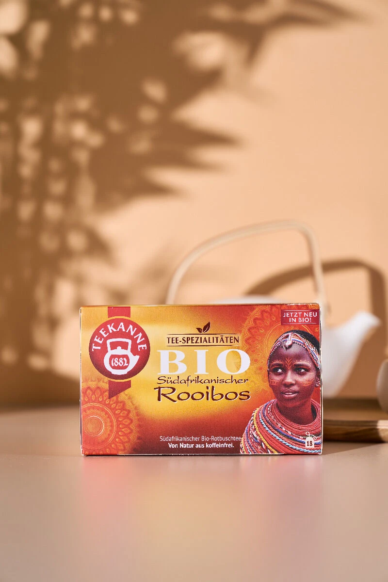 Eine Schachtel Südafrikanischer BIO Rooibos RFA-Tee steht auf einem Tisch und zeigt das Bild einer Frau in traditioneller Kleidung. Im Hintergrund erscheint eine unscharfe weiße Teekanne. Warmes Licht und Schatten von Blättern werfen Muster an die Wand.
