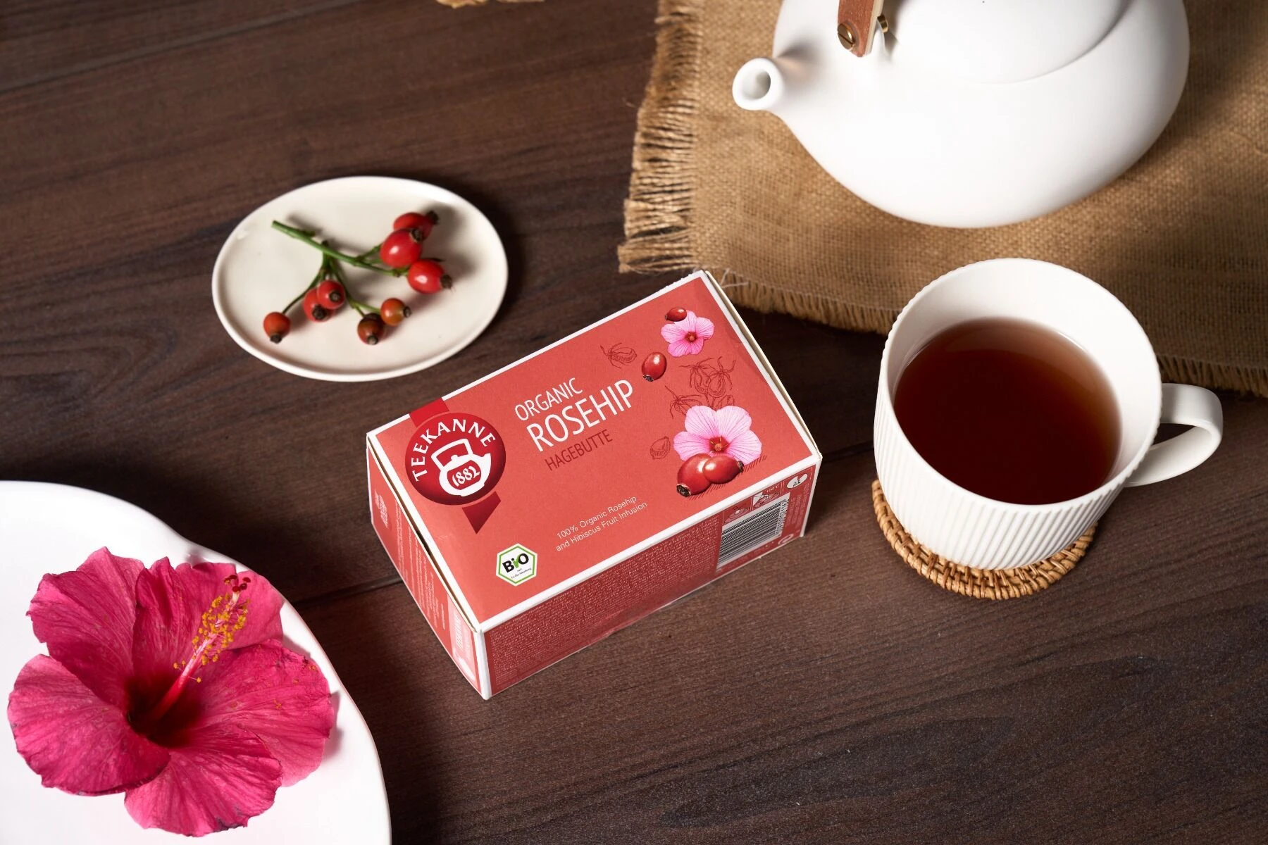 Eine Schachtel Bio-Hagebuttentee steht auf einem Tisch mit einer weißen Teetasse, einer Teekanne, frischen Hagebutten und einer rosafarbenen Hibiskusblüte, alles auf einem Tischset aus Sackleinen, das einen gemütlichen, einladenden Eindruck macht.
