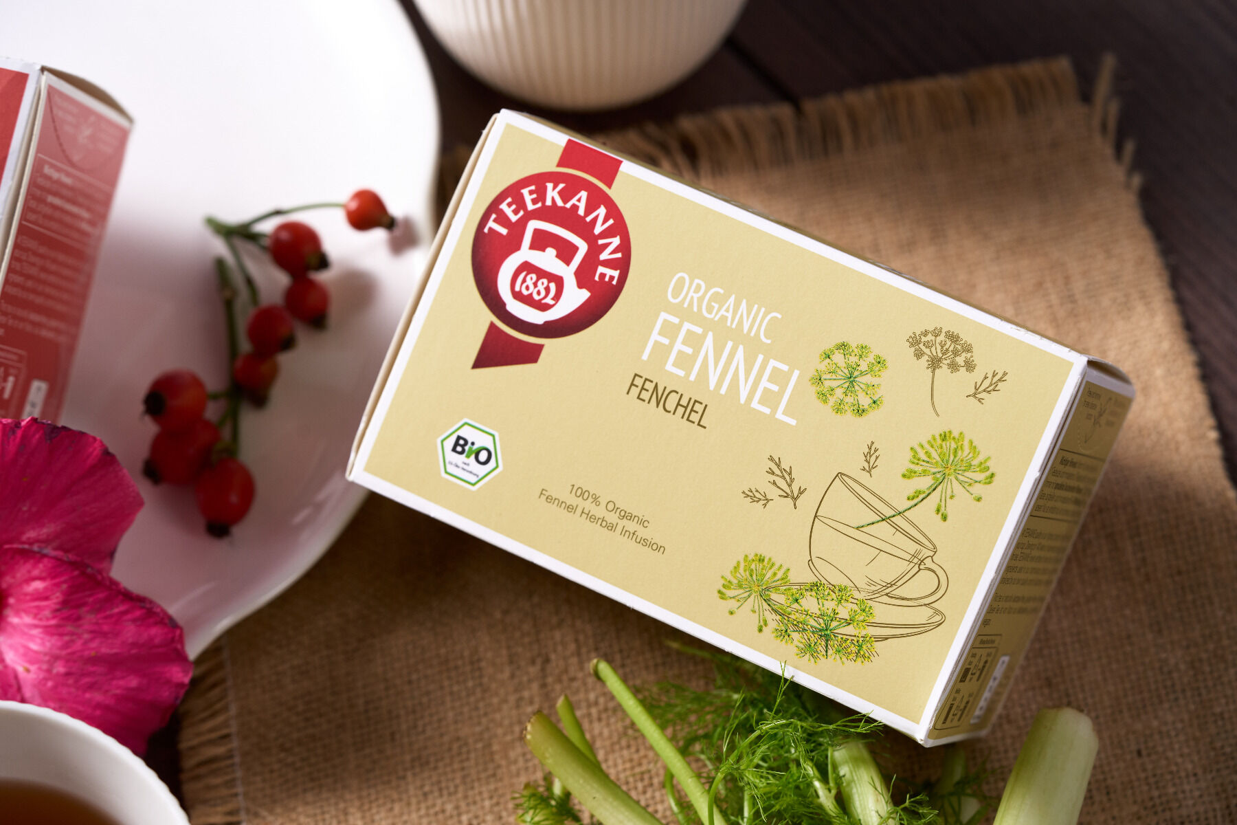 Eine Schachtel Bio-Fenchel von Teekanne liegt auf einer gewebten Matte, umgeben von Fenchel, einer roten Blüte, roten Beeren und einer weißen Tasse. Auf der Verpackung sind Fenchel-Illustrationen und eine dampfende Teetasse abgebildet.