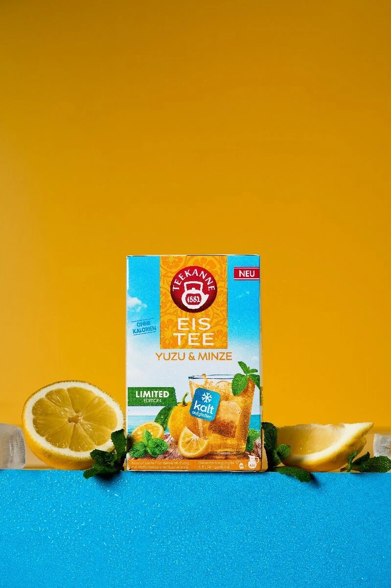 Eine Dose Yuzu & Minze liegt auf einer blauen Fläche mit Minzblättern, Eiswürfeln und Zitronenscheiben vor einem leuchtend gelben Hintergrund - perfekt zum kalt aufgießen und genießen.