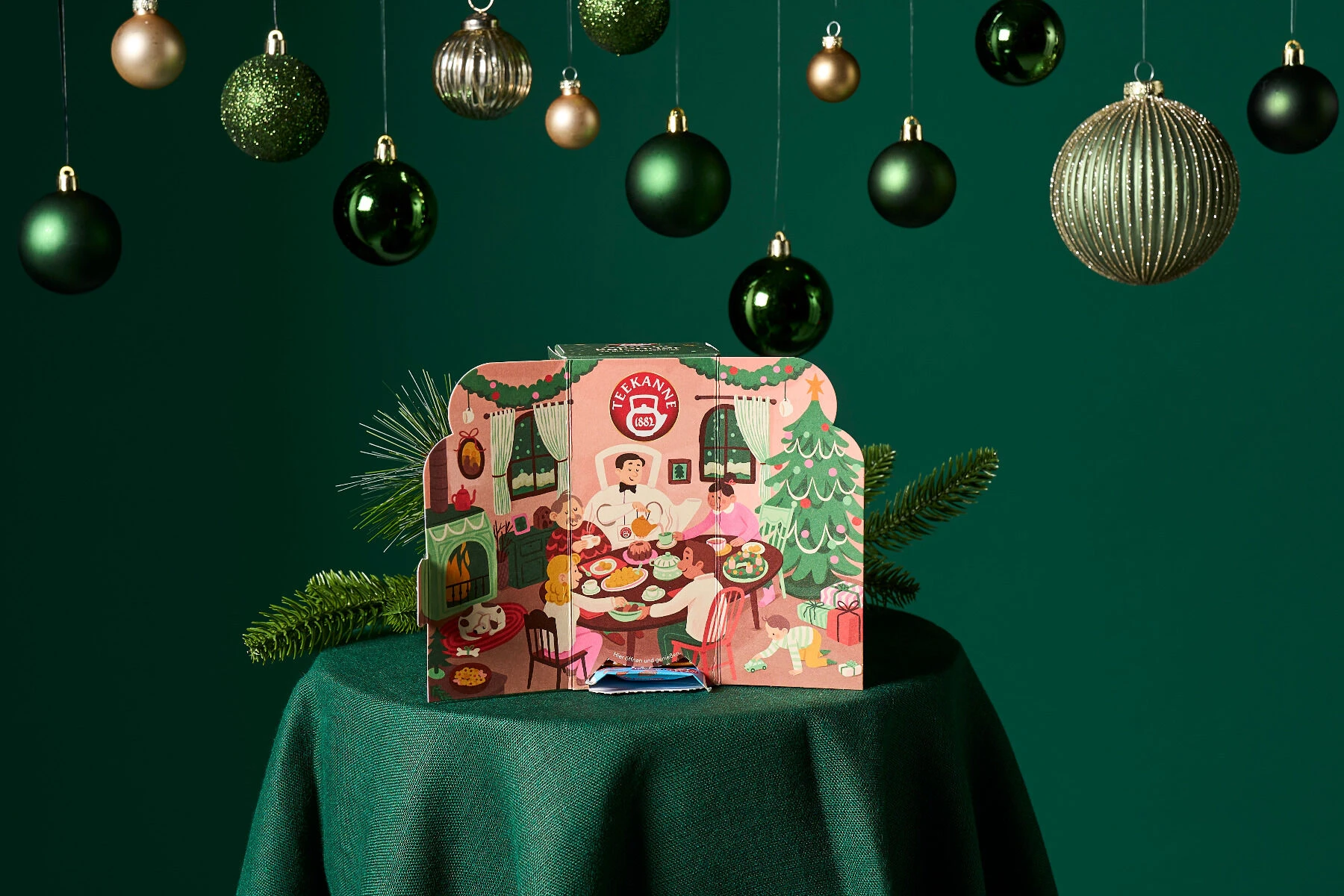 Die Pop-up-Karte Bio-Adventskalender zeigt eine festliche Szene mit einer Illustration eines Festessens auf einem grünen Tisch, umgeben von hängenden grünen und goldenen Ornamenten sowie Tannenzweigen.