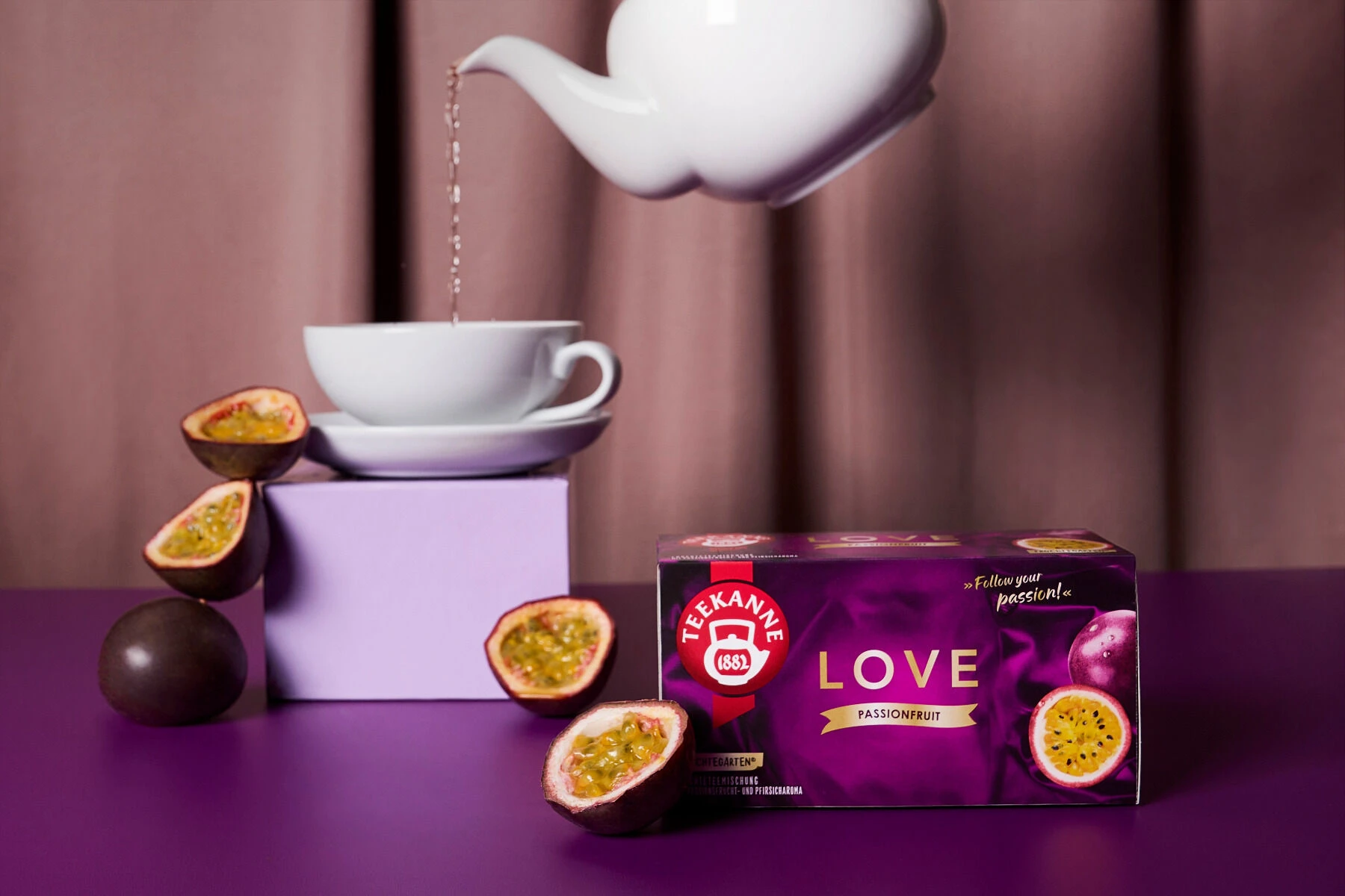 Aus einer weißen Teekanne wird Tee in eine Tasse gegossen, die neben frischen Passionsfrüchten und einer violetten Schachtel mit „LOVE Passionfruit“-Tee steht; alles ist auf einer violetten Unterlage angeordnet, vor dem Hintergrund eines mauvefarbenen Vor