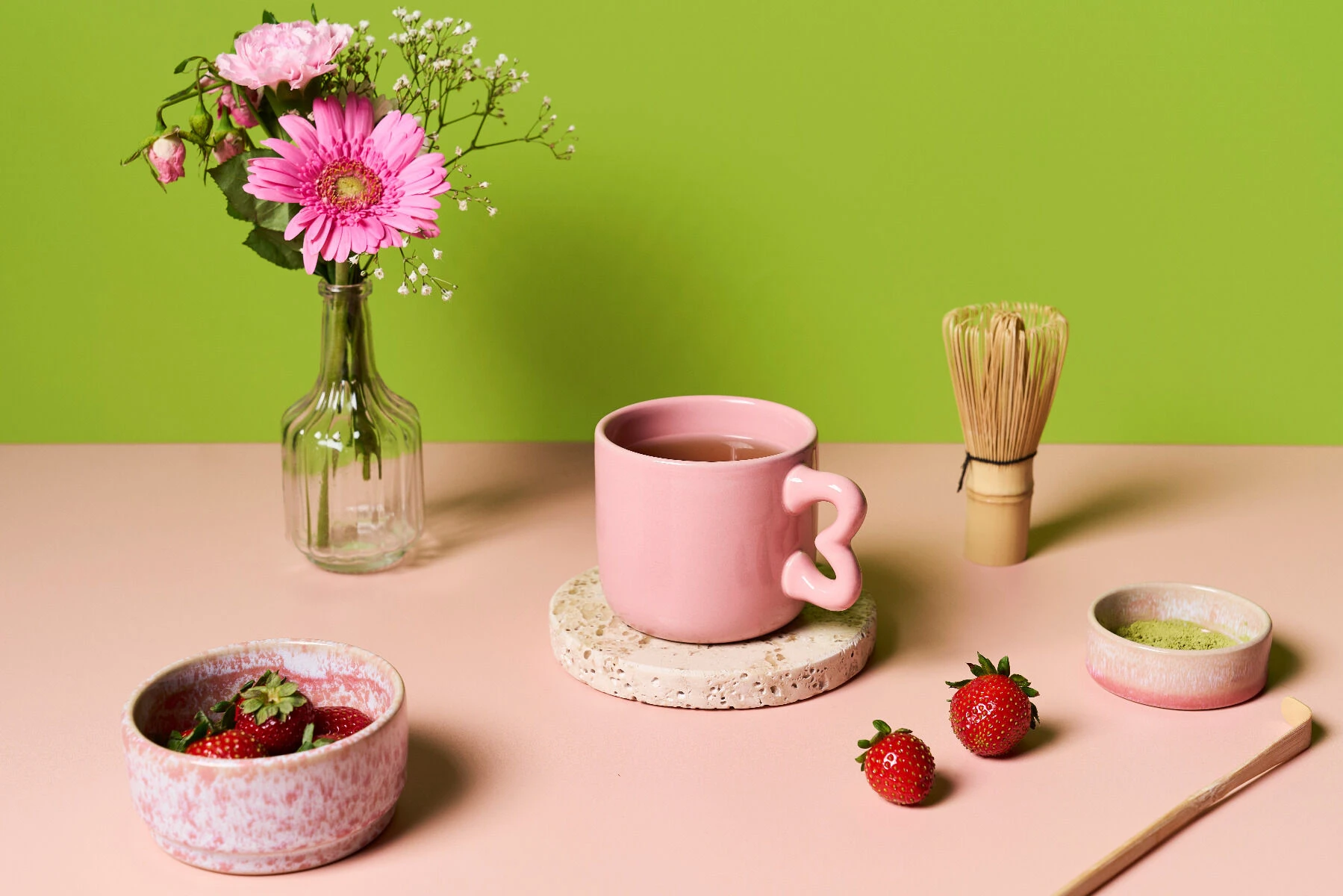 Eine rosa Tasse Tee steht auf einem Untersetzer, umgeben von Erdbeeren, einer Schale, „Grüner Tee Matcha Strawberry“, einem Bambusbesen und einer Vase mit rosa Blumen – alles auf einer rosa Fläche vor grünem Hintergrund angeordnet.