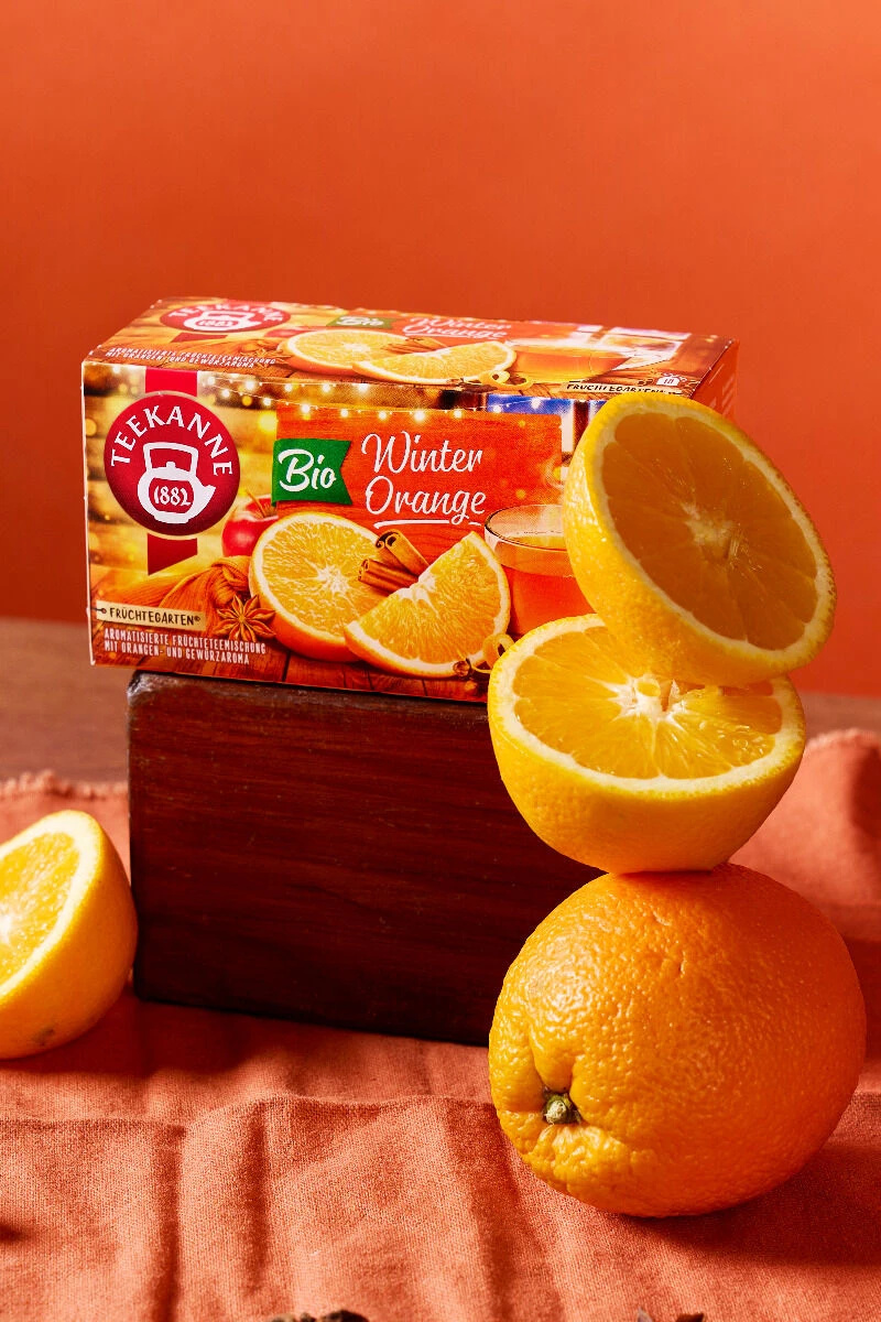 Eine Schachtel BIO-Winter-Orangentee wird zusammen mit frischen Orangen – sowohl ganzen als auch in Scheiben geschnittenen – auf einem orangefarbenen Tuch mit einem Holzblock vor einem orangefarbenen Hintergrund präsentiert, was an die warmen, aromatische