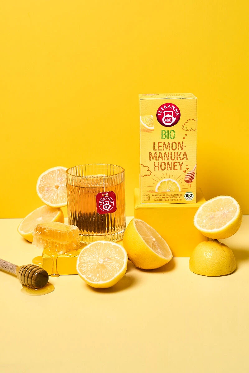 Ein Glas Tee, eine Honigwabe, Zitronenhälften und eine Schachtel mit der Aufschrift BIO Lemon Manuka Honey sind auf einer gelben Fläche vor einem passenden gelben Hintergrund angeordnet. Neben der Honigwabe liegt ein Honiglöffel.