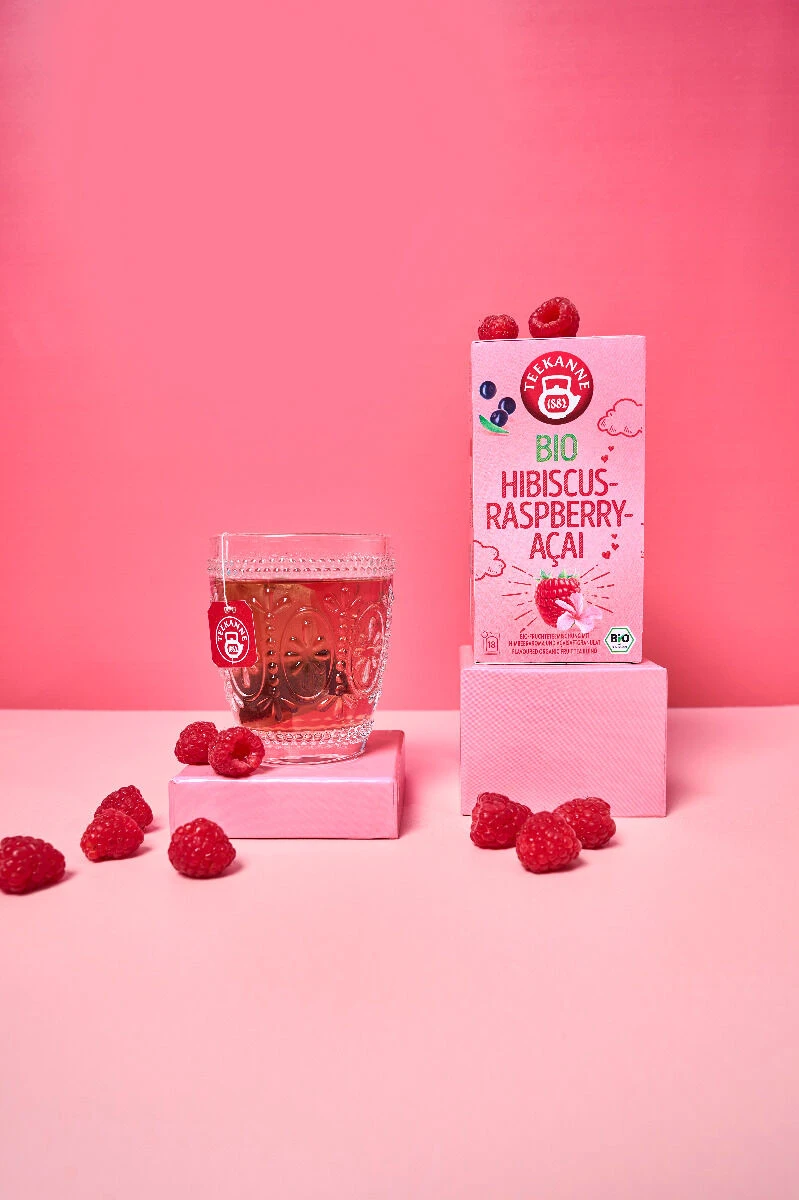 Ein Glas BIO-Hibiskus-Himbeer-Açaí-Tee neben der Produktverpackung, umgeben von frischen Himbeeren auf rosa Hintergrund.