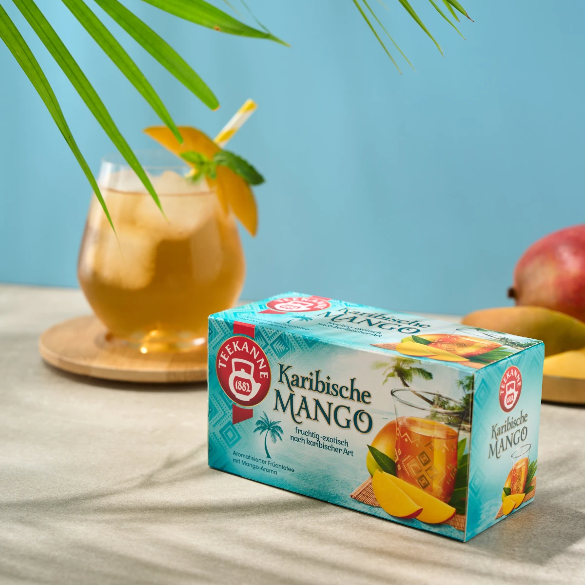 Auf einem Tisch steht eine Schachtel Karibische Mango, dahinter ein Glas Eistee, garniert mit Mango, und ein Schirmchen, umgeben von frischen Mangos und grünen Blättern vor einem hellblauen Hintergrund.