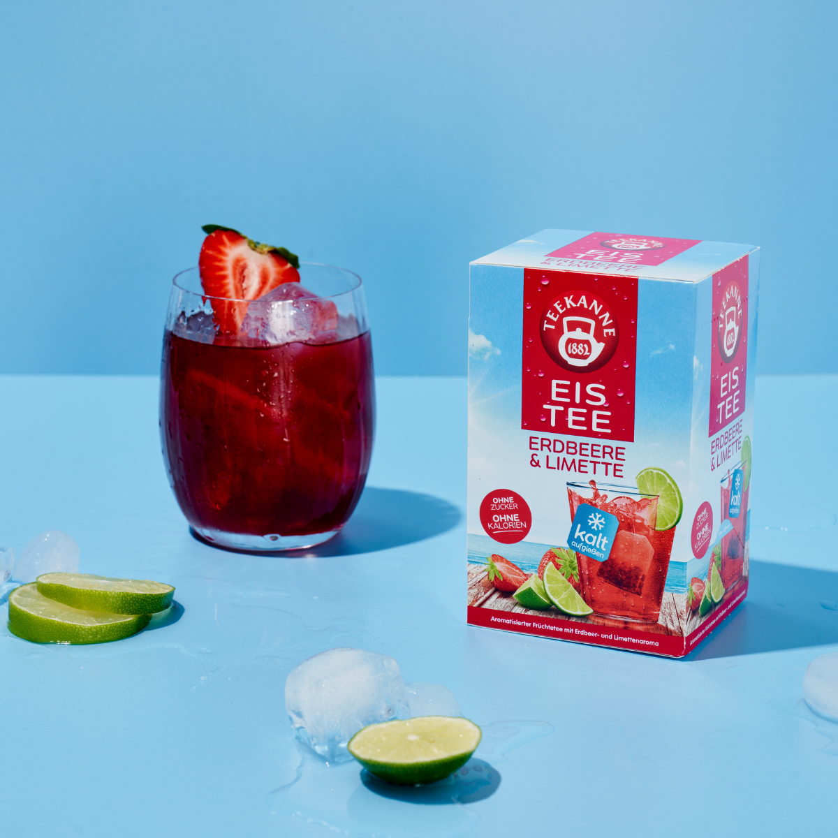 Ein Glas roter Eistee mit Eis und Erdbeergarnitur steht neben einer Schachtel Teekanne Cool Sensations Erdbeere & Limette. Eiswürfel und Limettenspalten sind auf der blauen Oberfläche verstreut.