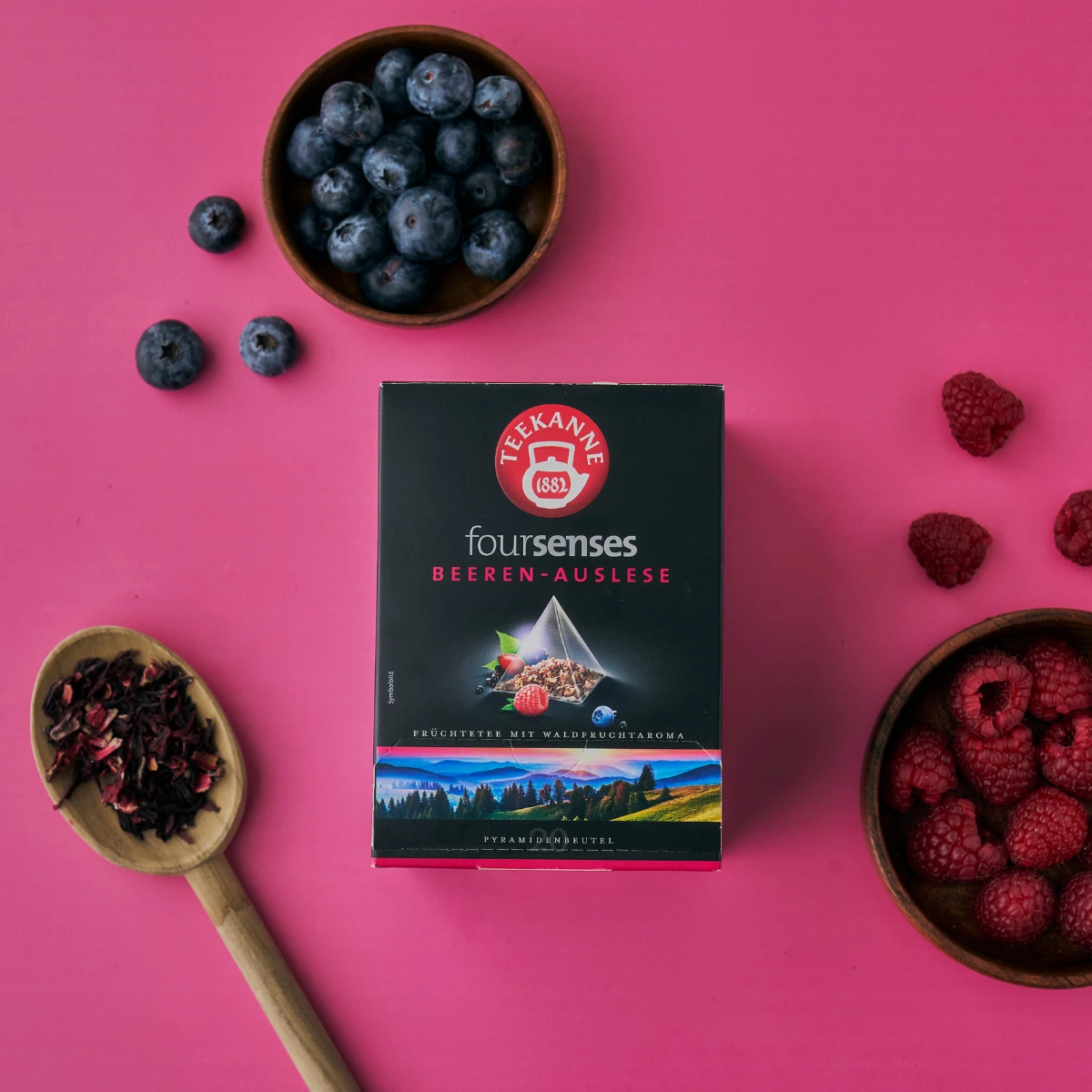 Eine Schachtel Beeren-Auslese-Tee wird zwischen frischen Blaubeeren, Himbeeren und einem Holzlöffel mit getrockneten Teeblättern auf einem leuchtend rosa Hintergrund präsentiert.