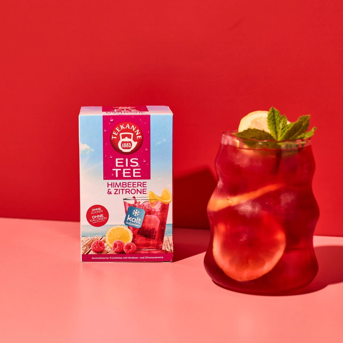 Eine Schachtel Himbeer-Zitronen-Tee steht neben einem Glas roten Eistees, garniert mit Zitronenscheiben und Minze, alles auf einer rosa Fläche vor einem roten Hintergrund.