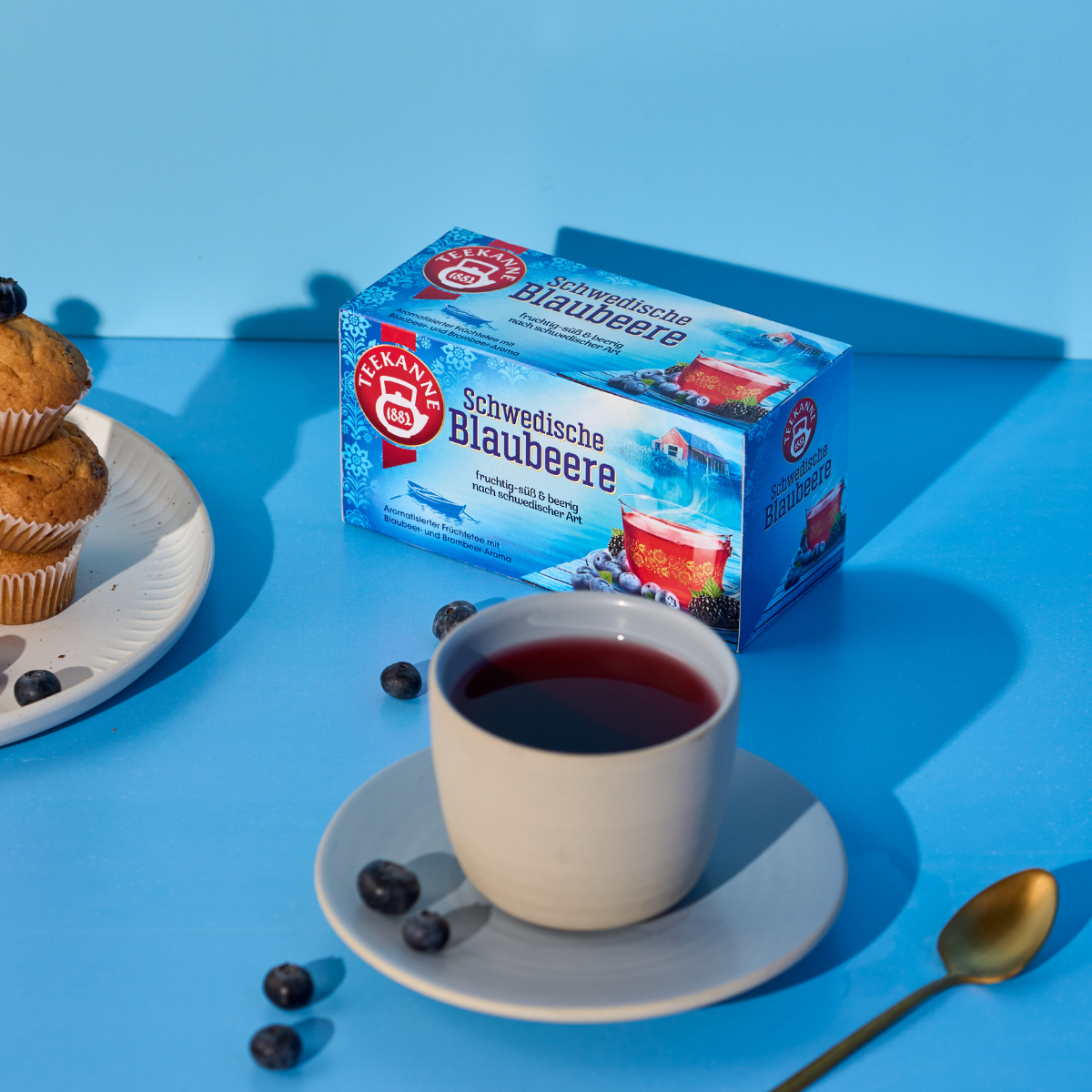 Eine Tasse Beerentee steht auf einer Untertasse neben verstreuten Blaubeeren, zwei Blaubeermuffins auf einem Teller und einer Schachtel Schwedischer Blaubeeren, alles auf einem blauen Hintergrund angeordnet.