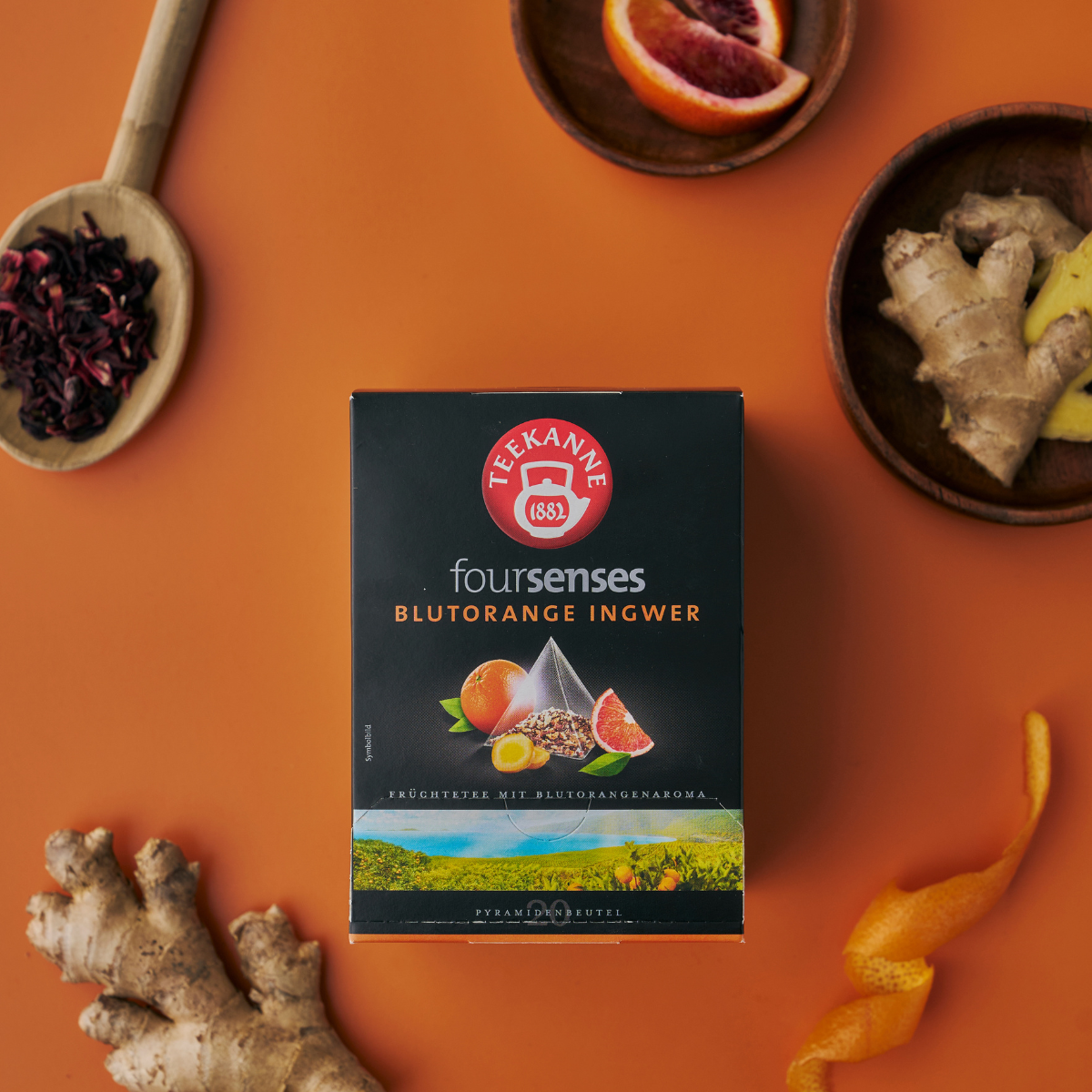 Eine Schachtel Blutorange-Ingwer-Tee steht auf einer orangefarbenen Oberfläche, umgeben von frischem Ingwer, Blutorangenscheiben, Hibiskusblütenblättern und Orangenschale.