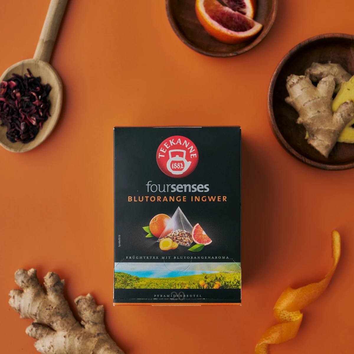 Eine Schachtel Blutorange-Ingwer-Tee steht auf einer orangefarbenen Oberfläche, umgeben von frischem Ingwer, Blutorangenscheiben, Hibiskusblütenblättern und Orangenschale.