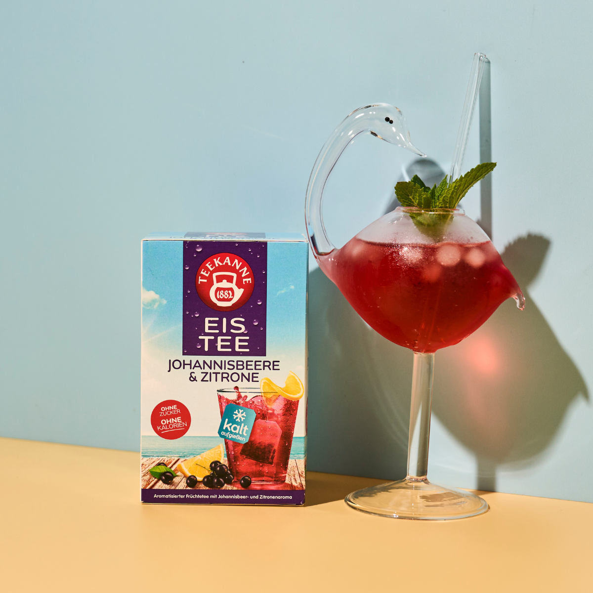 Eine Schachtel Johannisbeere-Zitrone steht neben einem schwanenförmigen Glas, gefüllt mit kaltem Tee, Eiswürfeln, Beeren und Minzblättern, vor einem pastellgelben und blauen Hintergrund.
