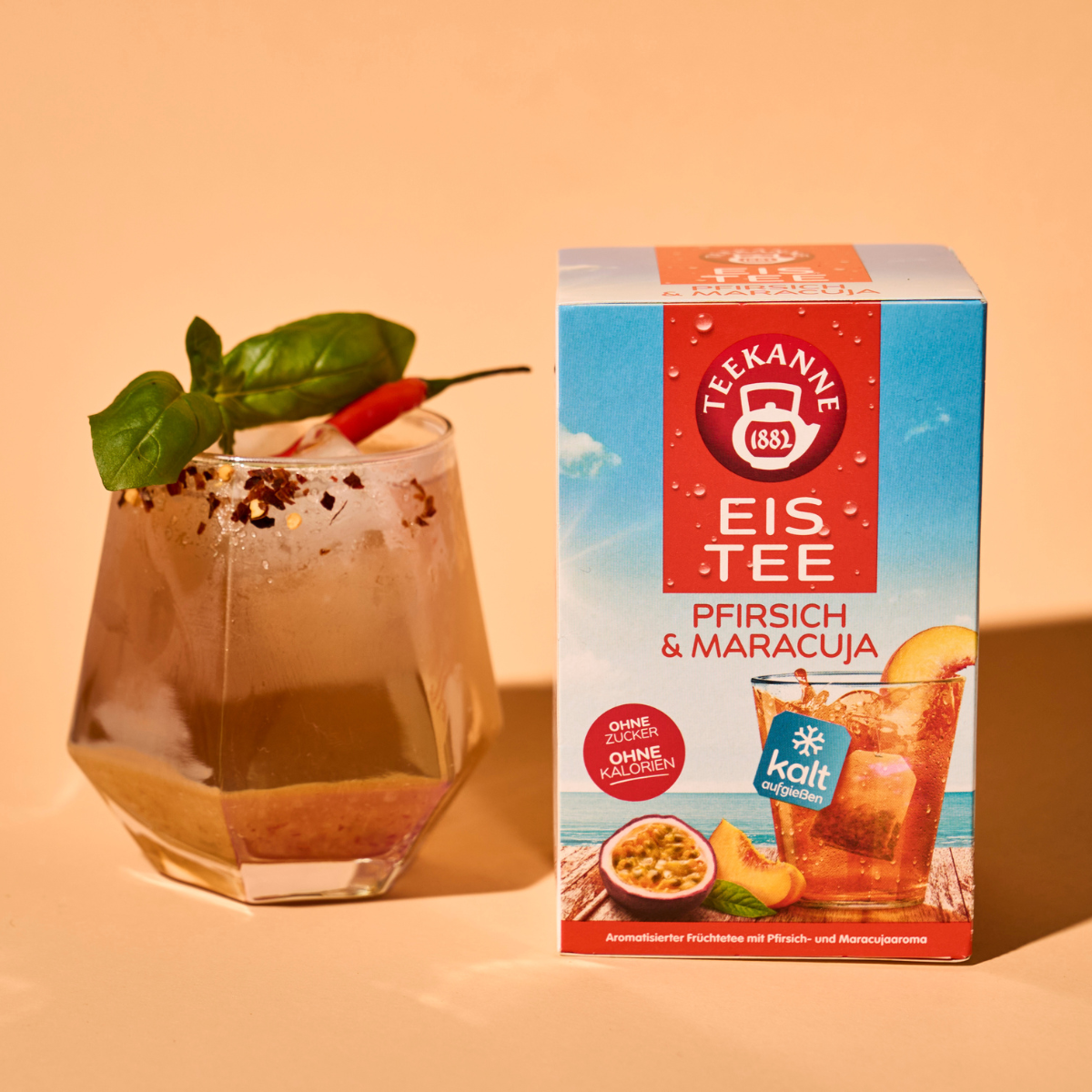 Ein Glas Eistee mit Basilikum und Chili steht neben einer Schachtel Pfirsich & Maracuja auf einer beigen Oberfläche und präsentiert seinen erfrischenden Pfirsich- und Passionsfruchtgeschmack.