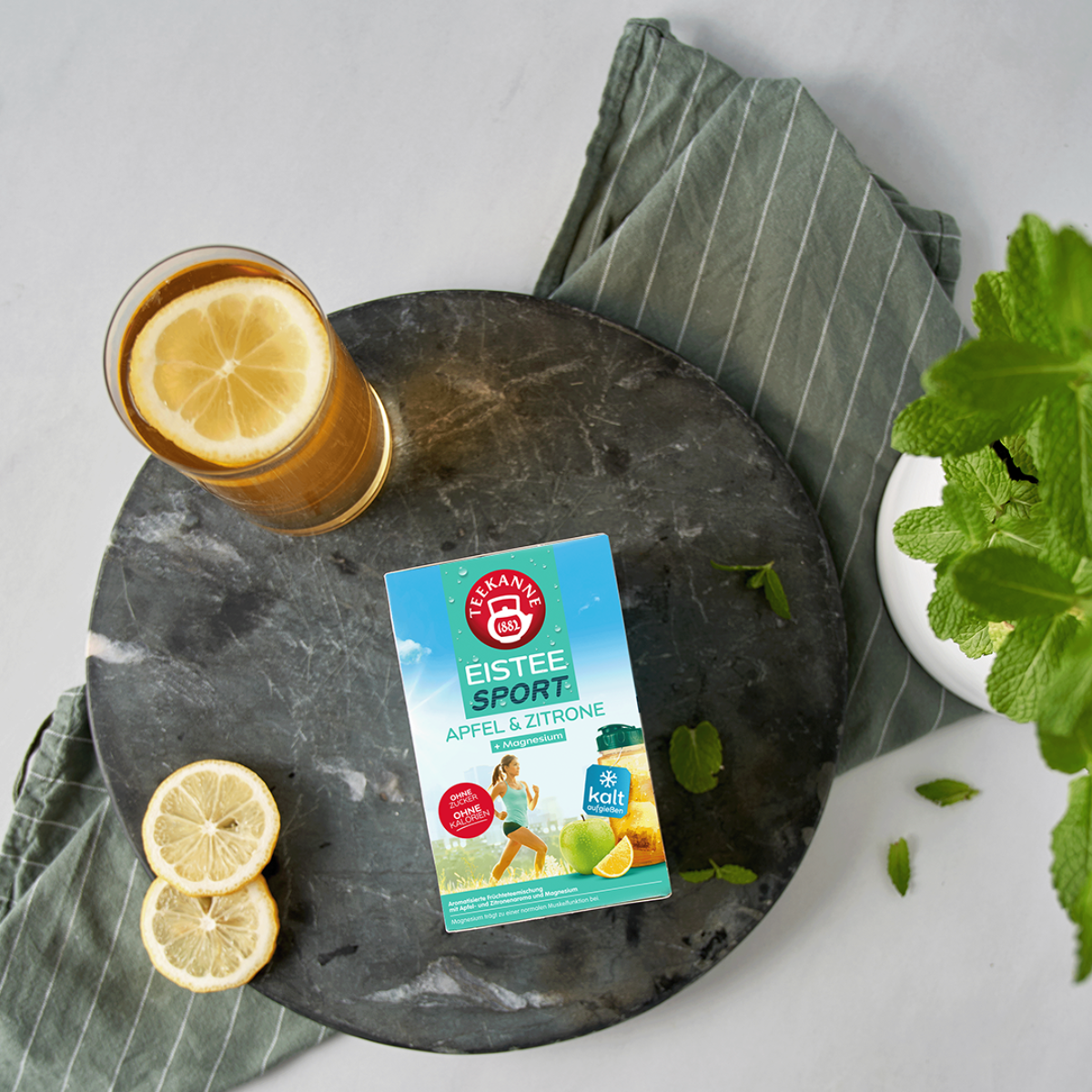 Ein Glas Eistee mit Zitrone, frischer Minze und die EISTEE SPORT APFEL-ZITRONE + Magnesium-Box stehen auf einem dunklen, runden Tablett über einem gestreiften Handtuch.