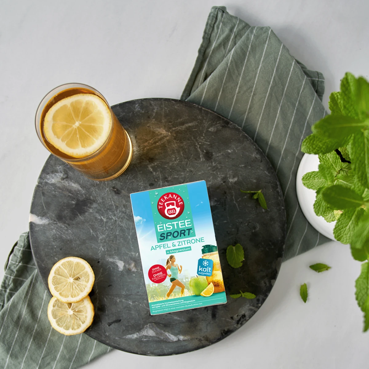 Ein Glas Eistee mit Zitrone, frischer Minze und die EISTEE SPORT APFEL-ZITRONE + Magnesium-Box stehen auf einem dunklen, runden Tablett über einem gestreiften Handtuch.