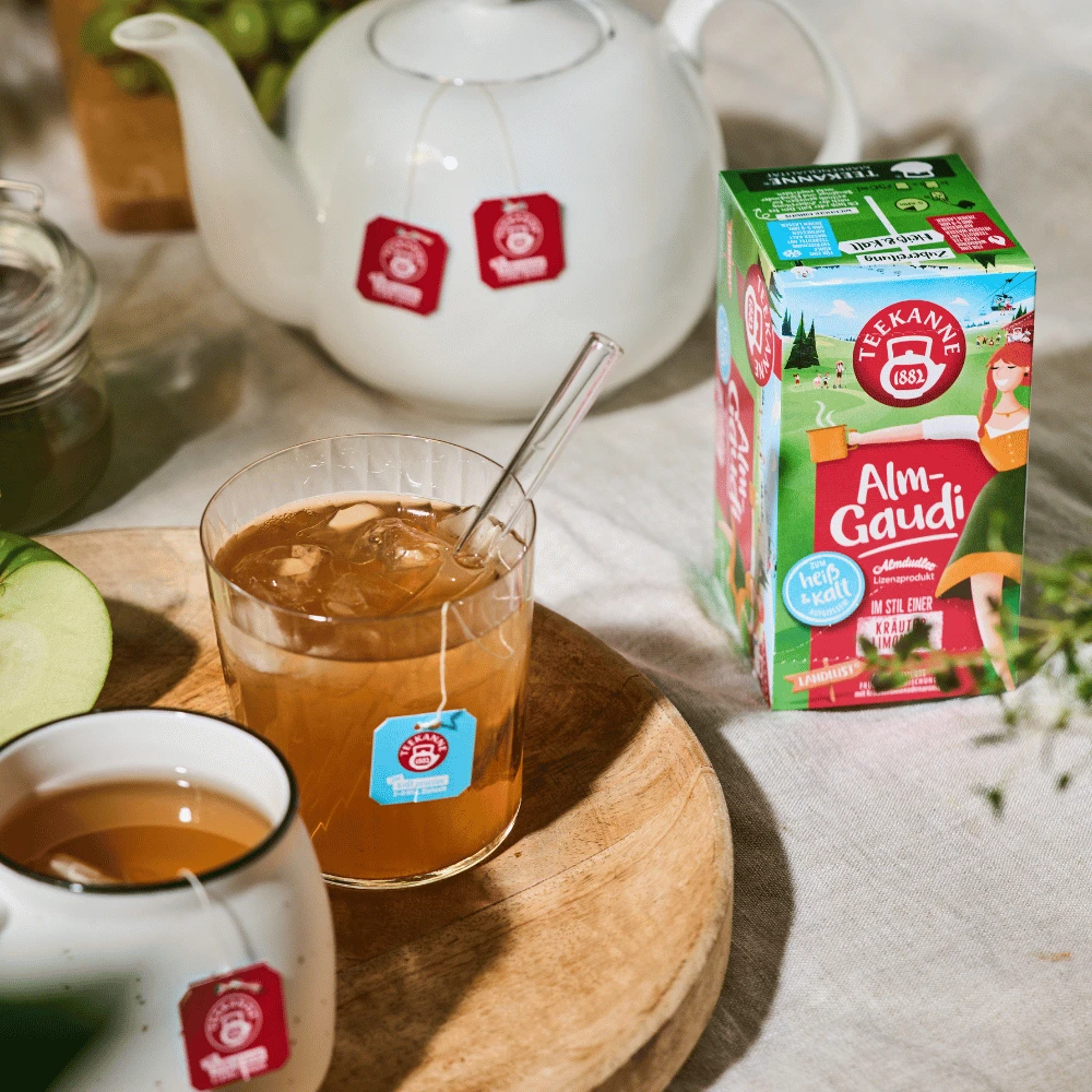 Ein Glas Eistee mit Strohhalm und eine Tasse heißer Tee stehen auf einem Holztablett neben einer weißen Teekanne und der bunten Almgaudi-Teebox. Grüne Apfelspalten, Weintrauben und erfrischende Kräuterlimonade ergänzen das einladende Bild.