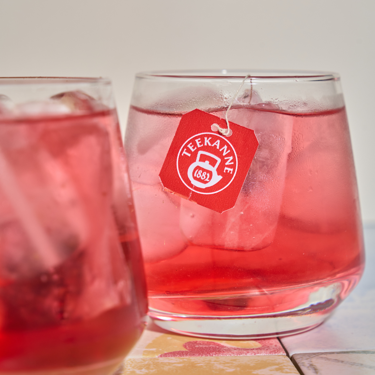 Zwei Gläser Eistee Mix & Match, jeweils mit Eiswürfeln und einem sichtbaren Teekanne-Teebeutelanhänger über dem Rand, versprechen erfrischende Kühle vor hellem Hintergrund.