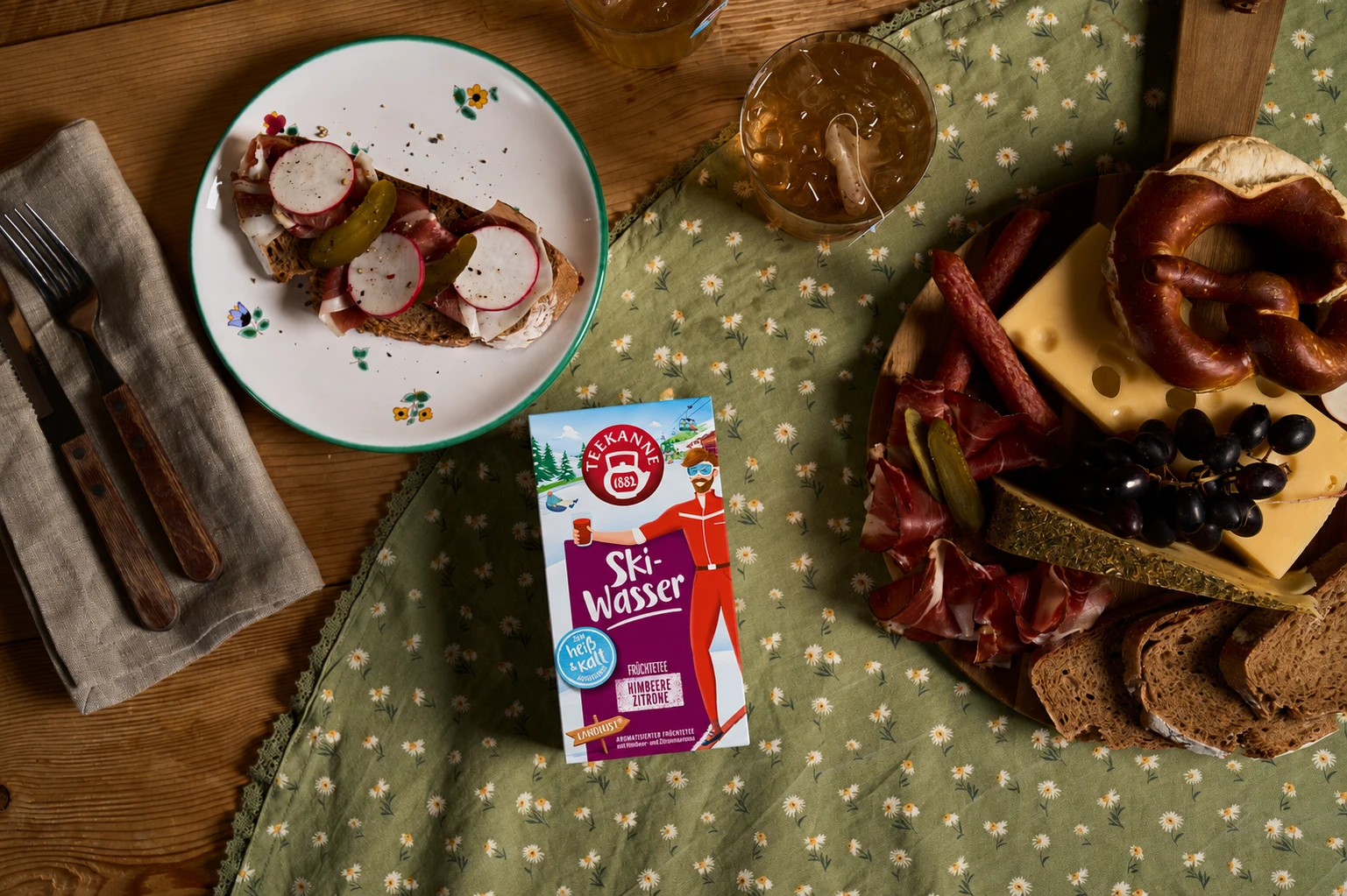 Auf einem Holztisch stehen offene Sandwiches, eine Käse- und Wurstplatte, Brot, eine Brezel und zwei Getränke – eines davon aus „fruchtiger Tee zum Kaltaufgießen“ – sowie in der Mitte ein „Skiwasser“-Saftkarton. Die Platte steht auf einem grünen Tuch mit 