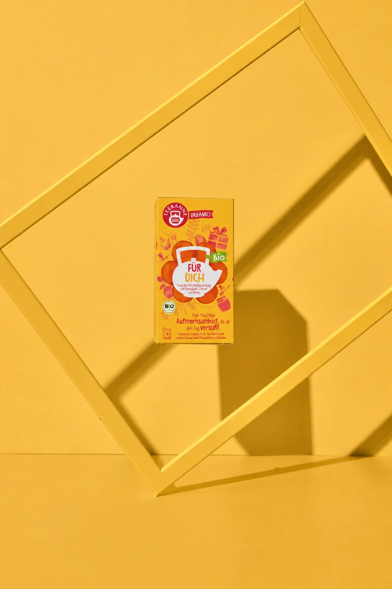 Eine Schachtel Organics „Für Dich“ Bio steht aufrecht auf einem leuchtend gelben Hintergrund, verziert mit orangefarbenen und floralen Motiven und eingerahmt von einem schräg stehenden gelben Quadrat – ein herrliches Bio-Früchtetee-Geschenk, perfekt für j