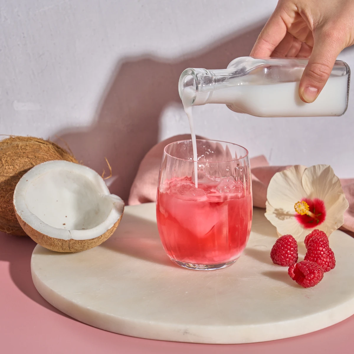 Eine Hand gießt Kokos & Hibiskus aus einer kleinen Glasflasche in ein rosa Eisgetränk, umgeben von Kokosnusshälften, Himbeeren und Hibiskus auf einer runden weißen Tafel – ein erfrischender zuckerfreier Sommergenuss.