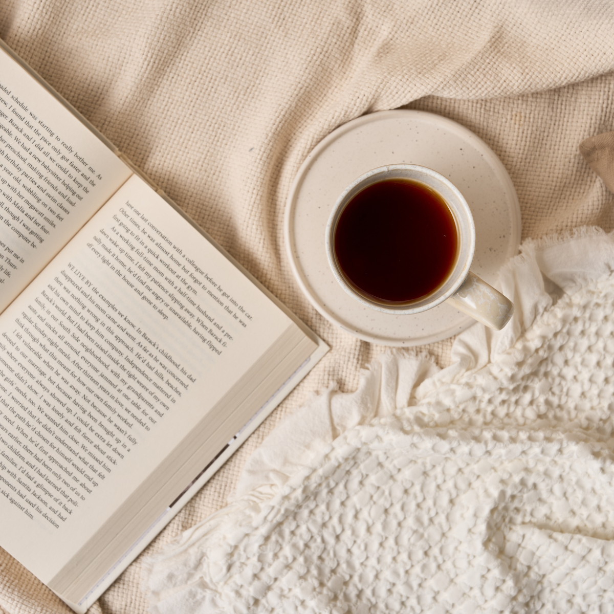 Eine Tasse schwarzen Kaffee auf einer Untertasse steht neben einem offenen Buch auf einer beigefarbenen strukturierten Decke und schafft eine gemütliche Atmosphäre, inspiriert von der BIO-Mischung „Innere Ruhe“.