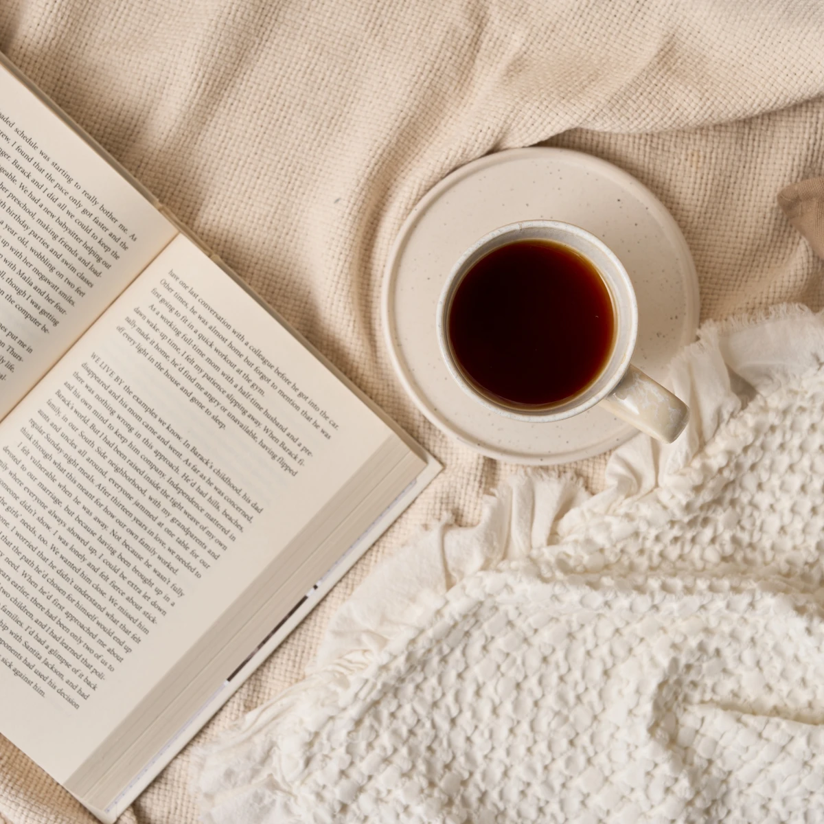 Eine Tasse schwarzen Kaffee auf einer Untertasse steht neben einem offenen Buch auf einer beigefarbenen strukturierten Decke und schafft eine gemütliche Atmosphäre, inspiriert von der BIO-Mischung „Innere Ruhe“.
