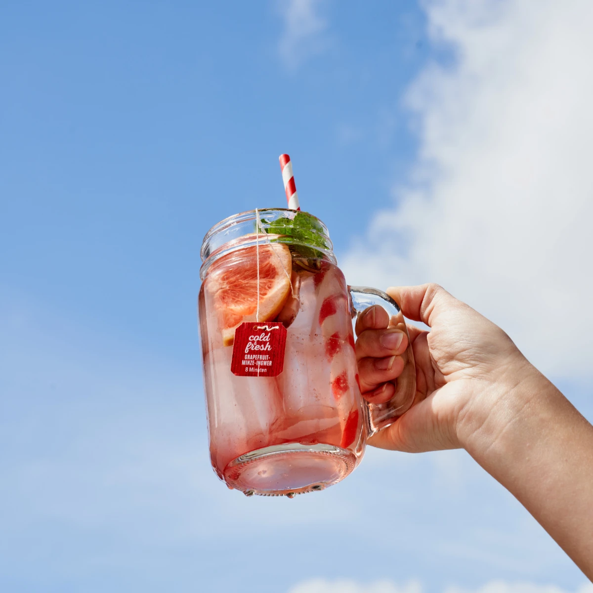 Eine Hand hält ein Einmachglas mit einem kalten und frischen Mix & Match Pink Grapefruit-Getränk, Eis, Grapefruitscheiben, Minze und einem gestreiften Strohhalm vor einem strahlend blauen Himmel hoch – die perfekte erfrischende Kombination.
