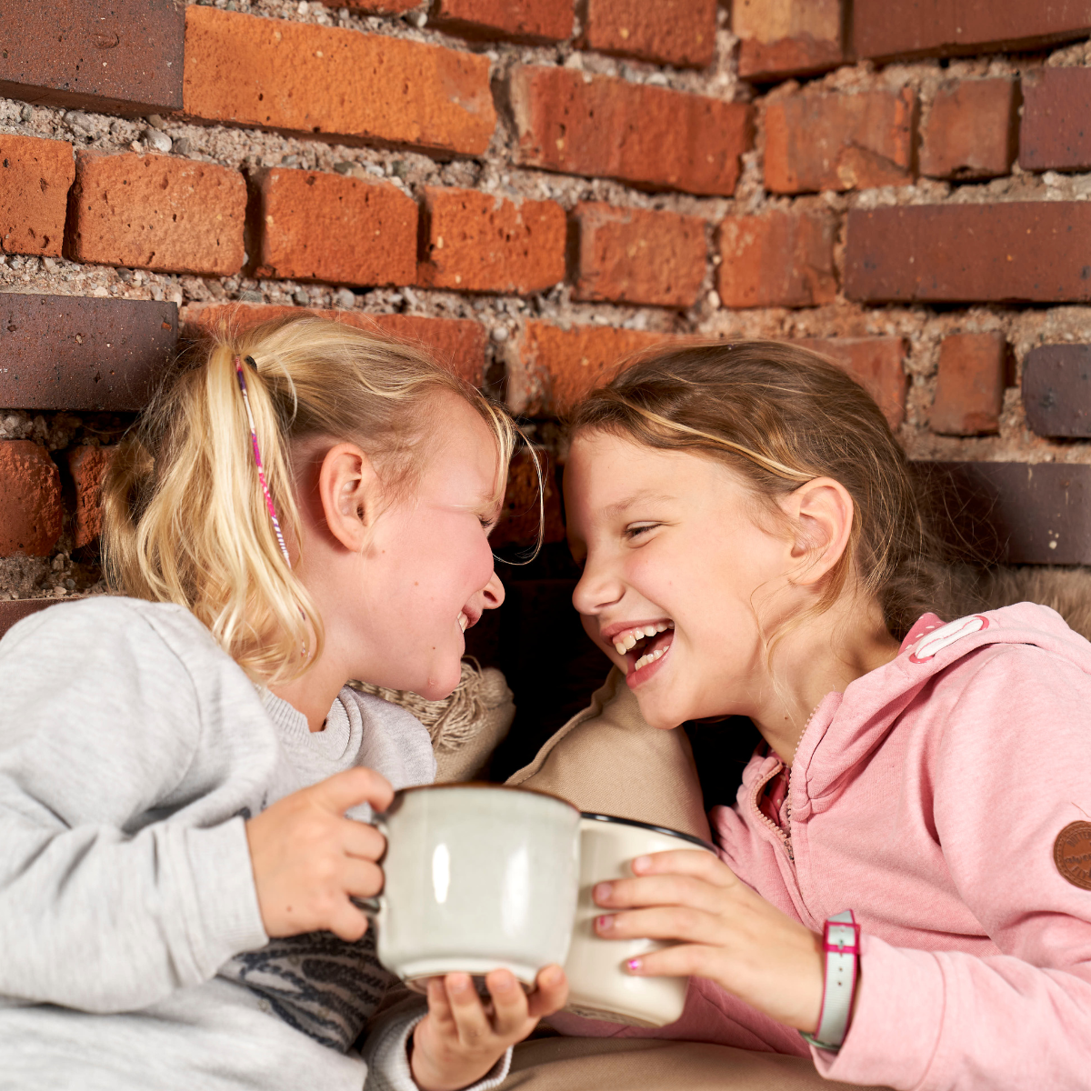 Zwei junge Mädchen sitzen dicht beieinander auf einem Sofa, lächeln und lachen und genießen dabei eine Tasse Bibo Beere Tee. Der gemütliche Moment wird durch eine Backsteinmauer im Hintergrund noch verstärkt.