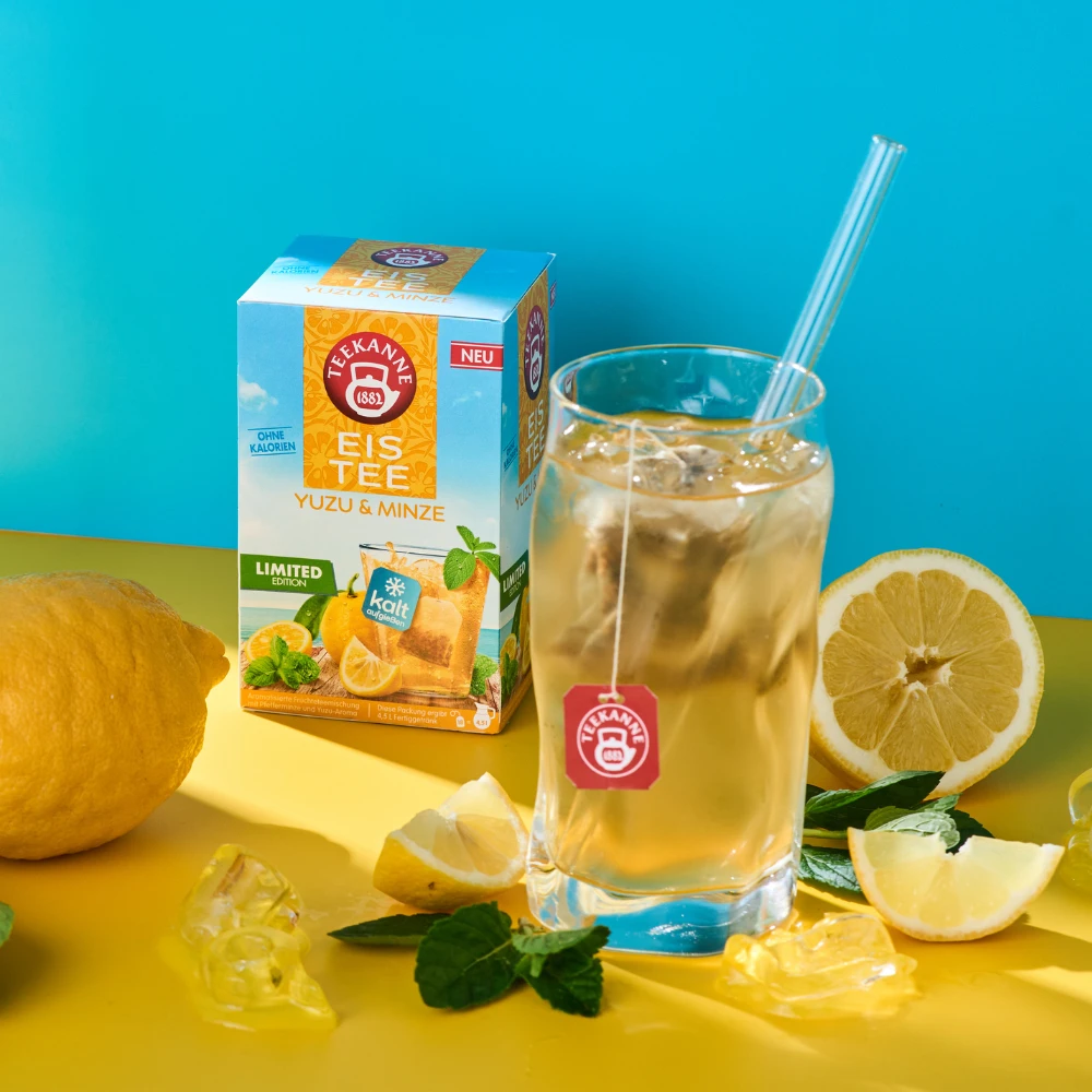 Ein Glas Eistee mit Strohhalm steht neben einer Schachtel Yuzu & Minze, umgeben von frischen Zitronen, Zitronenscheiben, Minzblättern und Eiswürfeln - ideal für den Genuss eines erfrischenden kalten Früchtetees vor einem leuchtend gelben und blauen Hinter