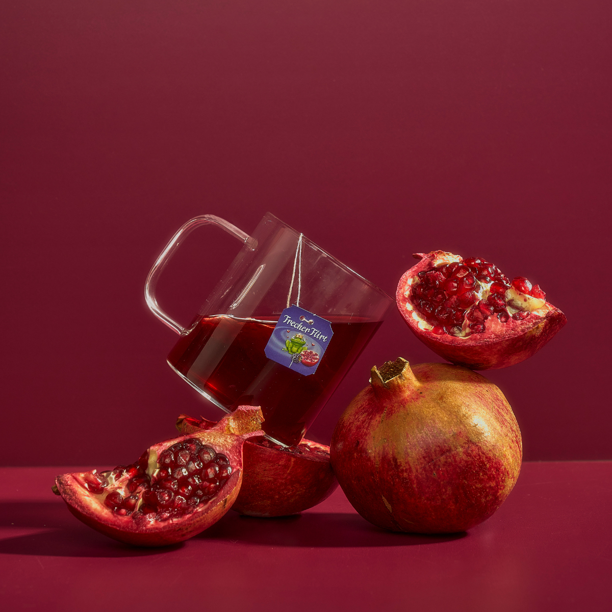 Eine Glastasse Frecher Flirt Granatapfeltee mit einem Teebeuteletikett steht zwischen ganzen und halbierten Granatäpfeln auf einem tiefroten Hintergrund.