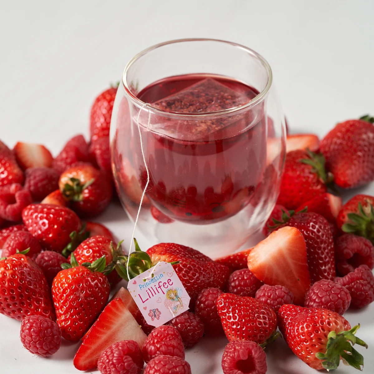 Ein doppelwandiges Glas Prinzessin Lillifee Tee ist von frischen Erdbeeren und Himbeeren umgeben. Ein Prinzessin Lillifee Teebeutel liegt daneben, alles auf einer makellos weißen Oberfläche.