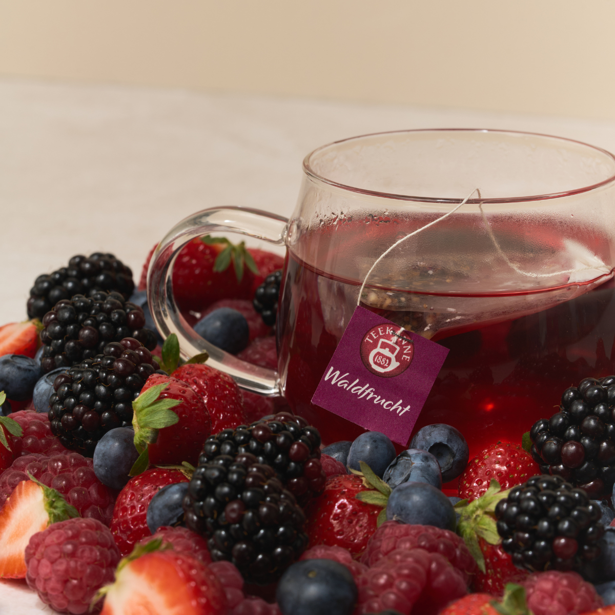Eine Glastasse mit tiefrotem Waldfruchttee steht zwischen frischen Erdbeeren, Brombeeren, Heidelbeeren und Himbeeren. Ein Teebeutel mit der Aufschrift „Waldfrucht“ hängt über dem Tassenrand.