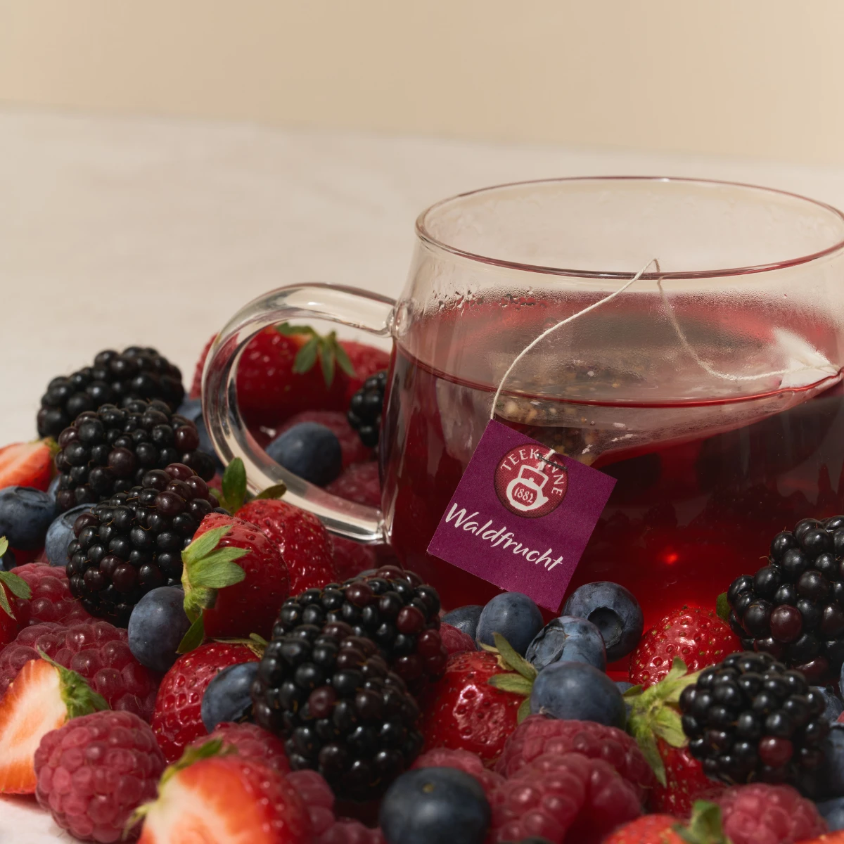 Eine Glastasse mit tiefrotem Waldfruchttee steht zwischen frischen Erdbeeren, Brombeeren, Heidelbeeren und Himbeeren. Ein Teebeutel mit der Aufschrift „Waldfrucht“ hängt über dem Tassenrand.