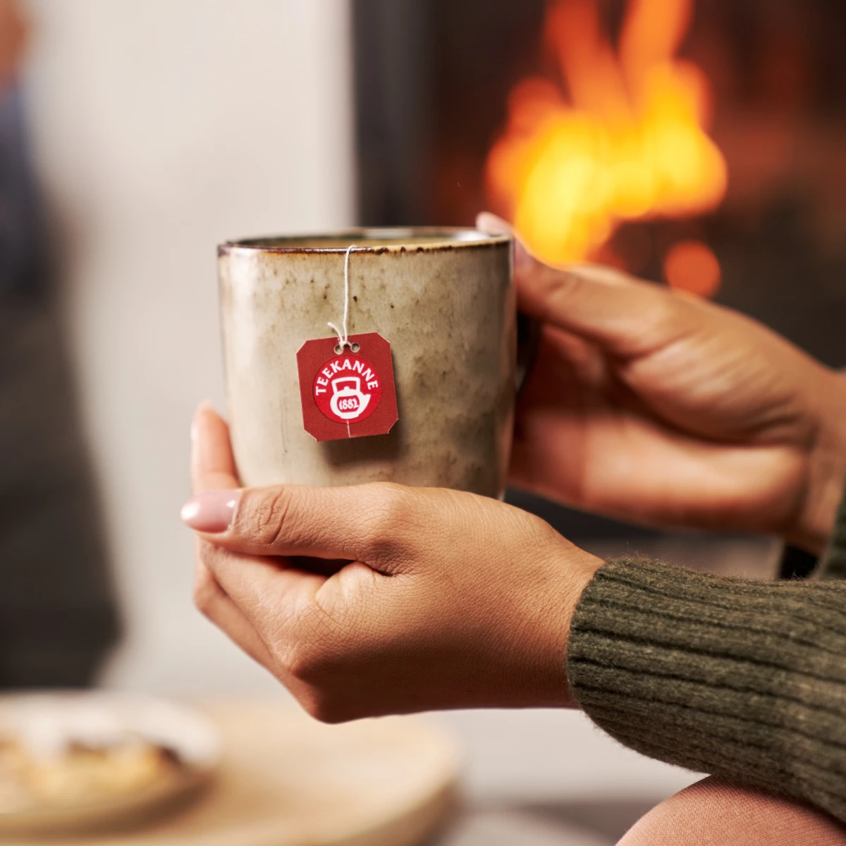 Eine Person hält eine Tasse ORGANICS Taste of Winter BIO vor einem warmen, verschwommenen Kaminfeuer. Ein Hauch von Apfel und Gewürzen erfüllt die gemütliche Atmosphäre und vermittelt Behaglichkeit und Entspannung – perfekt für einen wohltuenden Wintertee