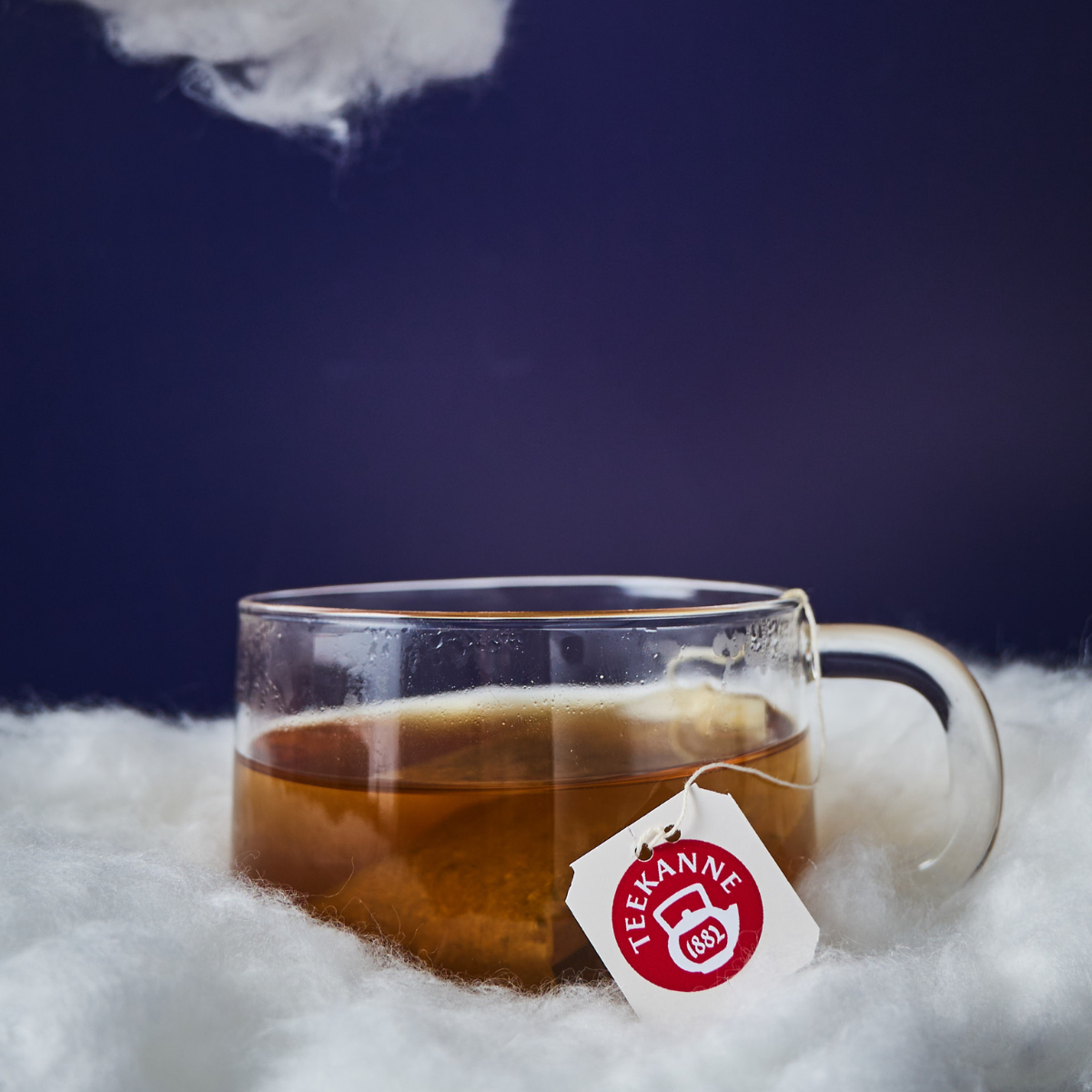 Eine klare Glastasse Tee mit einem Teekanne-Teebeutel steht auf einer flauschigen weißen Oberfläche wie Wolken, unter einem Fleck aus weißem Flaum und einem dunkelblauen Hintergrund – eine perfekte Kulisse, um „Besser Einschlafen“ zu genießen.