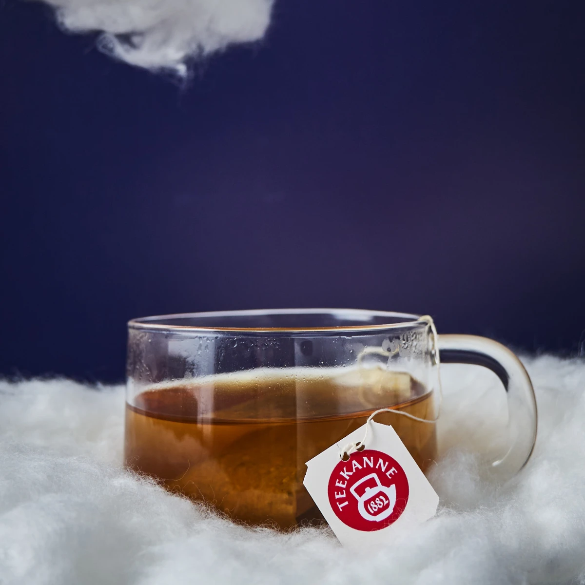 Eine klare Glastasse Tee mit einem Teekanne-Teebeutel steht auf einer flauschigen weißen Oberfläche wie Wolken, unter einem Fleck aus weißem Flaum und einem dunkelblauen Hintergrund – eine perfekte Kulisse, um „Besser Einschlafen“ zu genießen.