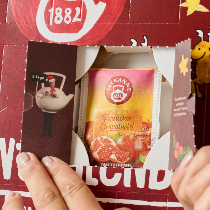 Jemand öffnet ein Türchen eines Adventskalenders und findet darin eine Packung Teekanne Granatapfel-Früchtetee. Der Adventskalender ist mit festlichen Motiven wie einem Weihnachtsmann und Rentieren verziert.
