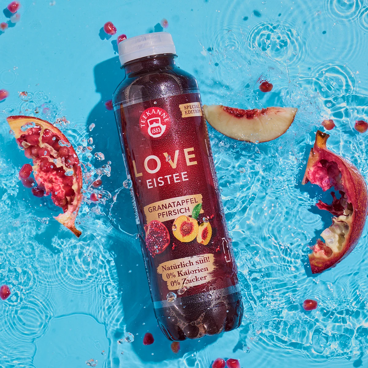 Eine Flasche Teekanne Granatapfel-Pfirsich-Eistee schwimmt auf blauem Wasser, umgeben von Spritzern, Früchten, Granatapfelkernen und Pfirsichscheiben.