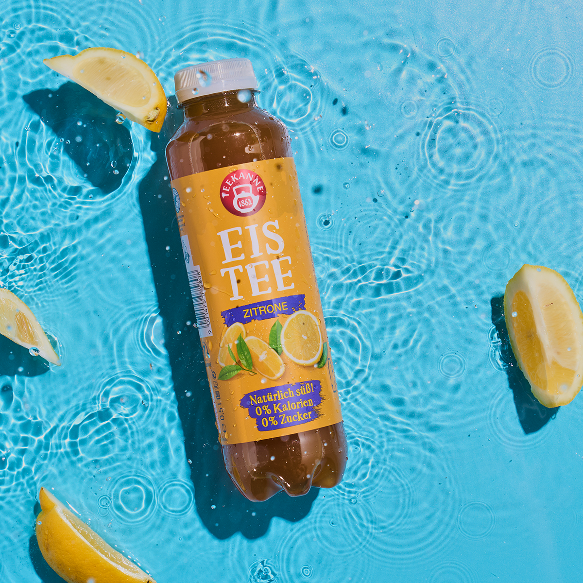 Eine Flasche Zitrone schwimmt in einem Becken mit plätscherndem Wasser, umgeben von Zitronenspalten. Das Etikett unterstreicht den natürlich süßen Schwarztee-Geschmack mit 0 % Kalorien und 0 % Zucker.