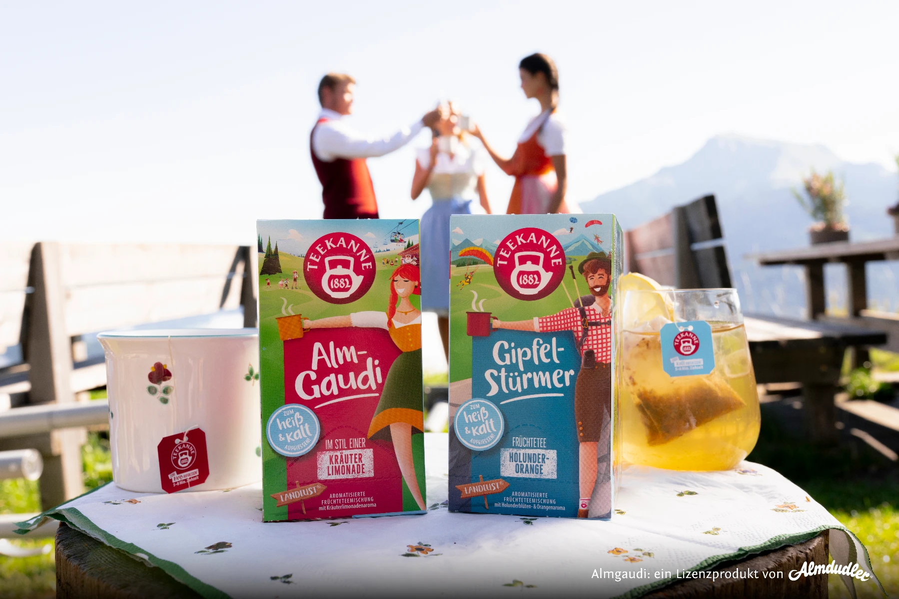 Zwei Schachteln Teekanne Gipfelstürmer-Tee, ein Glas Eistee und ein Becher stehen auf einem dekorierten Tisch im Freien. Im unscharfen Hintergrund stoßen zwei Personen mit den Bergen und dem blauen Himmel im Rücken auf ihre Getränke an.