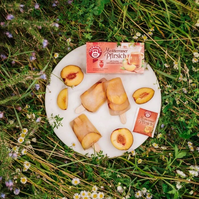 Drei Pfirsich-Eis am Stiel und in Scheiben geschnittene Pfirsiche sind auf einem runden weißen Teller in hohem Gras und Wildblumen arrangiert, daneben zwei Schachteln Mediterraner Pfirsich.