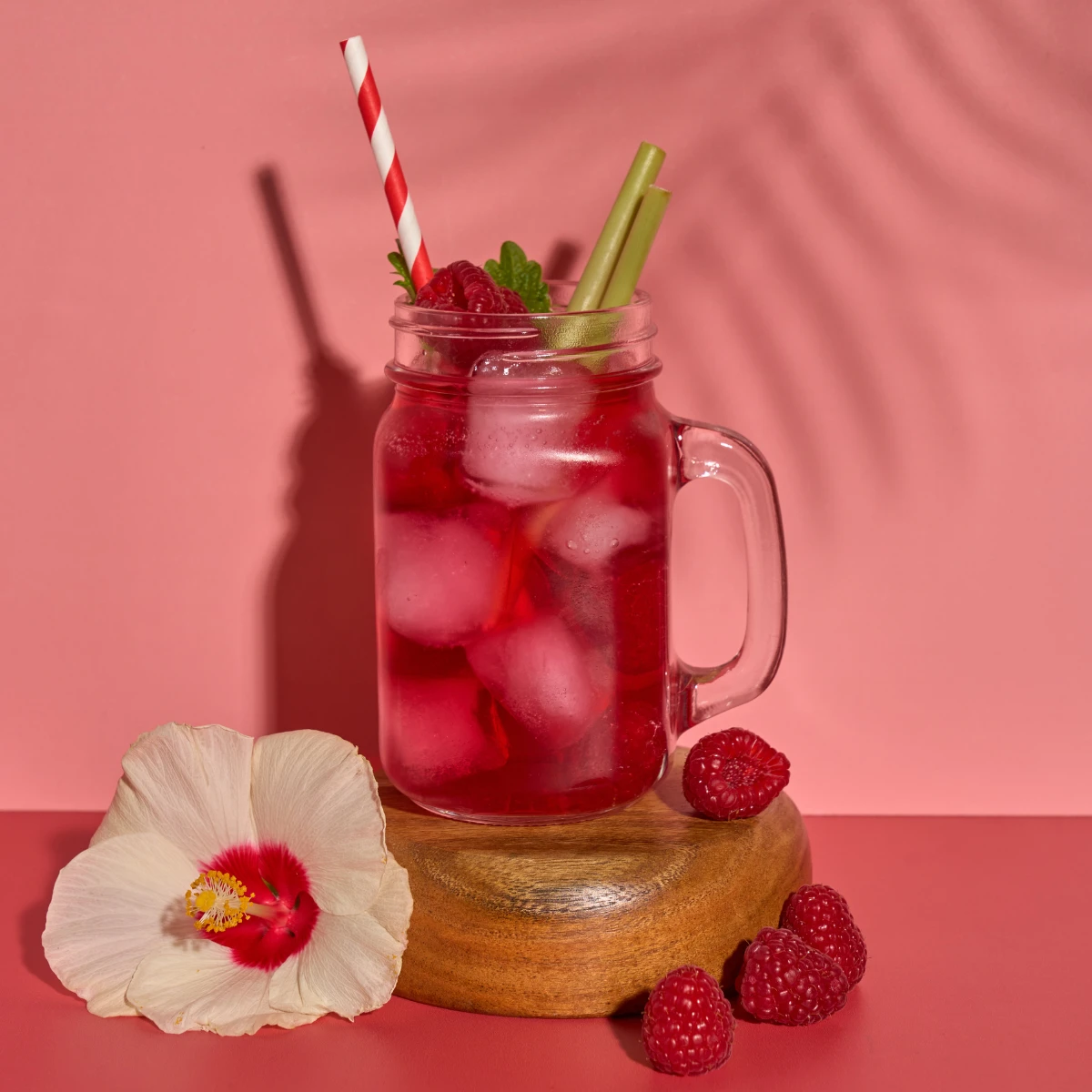 Ein Einmachglas mit kaltem und frischem Hibiskus-Himbeere-Zitronengras, gefüllt mit Eis und garniert mit Himbeeren, Minze und einem Strohhalm, steht auf einem Holzständer neben Hibiskusblüten und Himbeeren vor einem rosa Hintergrund.