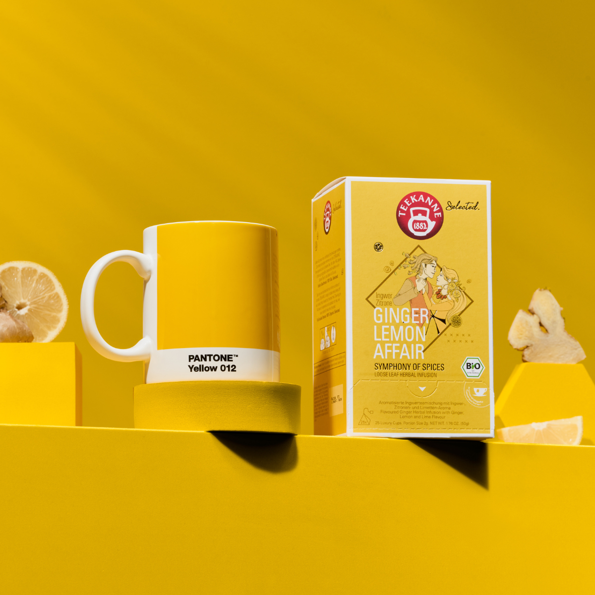 Eine Ginger Lemon Affair Luxury Cup BIO und ein gelber Pantone 012-Becher stehen auf einem gelben Hintergrund, akzentuiert mit Zitronenscheiben und Ingwerstücken.