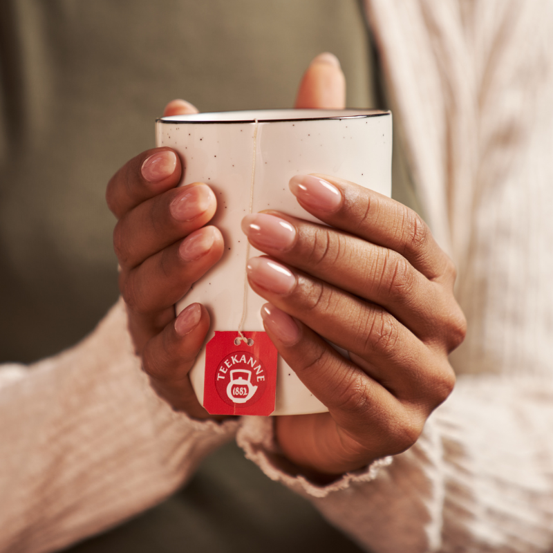 Eine Person mit sauber manikürten Nägeln hält eine weiß gesprenkelte Tasse mit der Aufschrift „Organics Mix & Match“-Tee und trägt einen leichten Strickpullover.