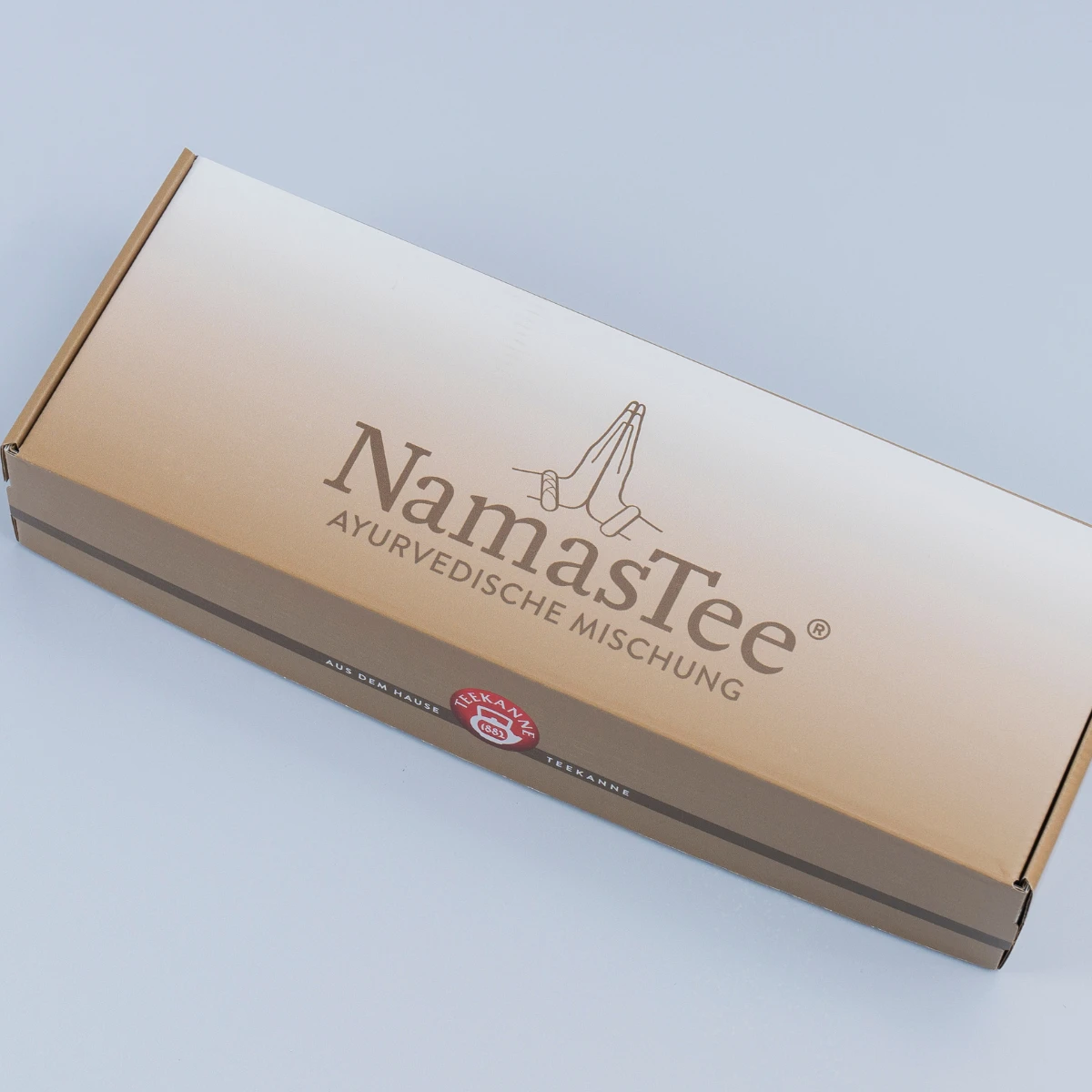 Eine rechteckige braune NamasTee-Geschenkbox mit der Aufschrift „AYURVEDISCHE MISCHUNG“ und einer Strichzeichnung betender Hände über dem Text, ideal als Bio-Geschenk, abgebildet auf hellgrauem Untergrund.