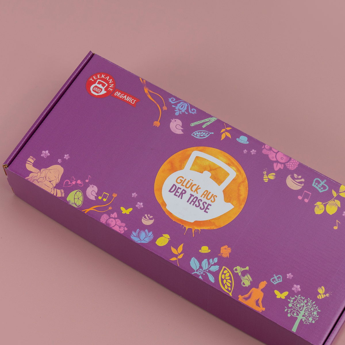 Eine violette, rechteckige Organics-Geschenkbox mit bunten Illustrationen und „Glück aus der Tasse“ in der Mitte auf rosa Hintergrund. Oben links befindet sich das rote Teekanne Organics Logo – ideal als Bio Kräutertee Geschenk.