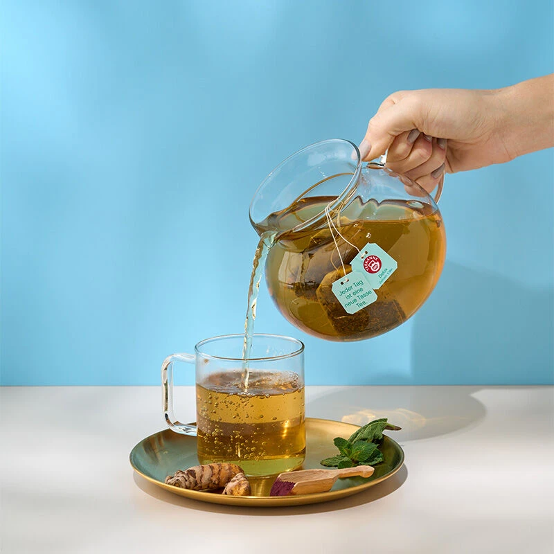 Eine Hand gießt Harmonietee Mix & Match aus einer Glasteekanne in eine klare Tasse auf einem goldenen Teller, umgeben von Ingwer, einem Honiglöffel und Minzblättern. Der hellblaue Hintergrund sorgt für einen ruhigen TEEKANNE-Teemoment.